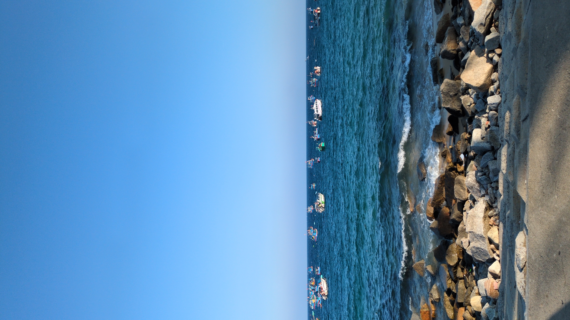 This image depicts a coastal scene in East Quy Nhon Ward, Vietnam, under a clear, bright blue sky. The upper approximately 40% of the frame shows an uninterrupted expanse of cloudless blue sky. The middle section, occupying approximately 40% of the image, features deep blue-green ocean water with moderate surface chop. Scattered across the visible water are numerous vessels, estimated to be around 40 in total. These include a predominance of smaller, multi-colored fishing boats, typically featuring white hulls with blue, green, and red accents, and at least two larger, predominantly white cabin cruiser-style boats. The lower approximately 20% of the image comprises the shoreline and immediate foreground. The shoreline consists of a natural or constructed rock barrier composed of various sized grey and brown stones, against which small waves are breaking. Pockets of tan sand are visible between the rocks at the waterline. The immediate foreground is a flat, light grey concrete surface, likely a pathway or promenade, with diagonal shadows cast upon it from an unseen object to the left. No individuals are visible in the frame.
