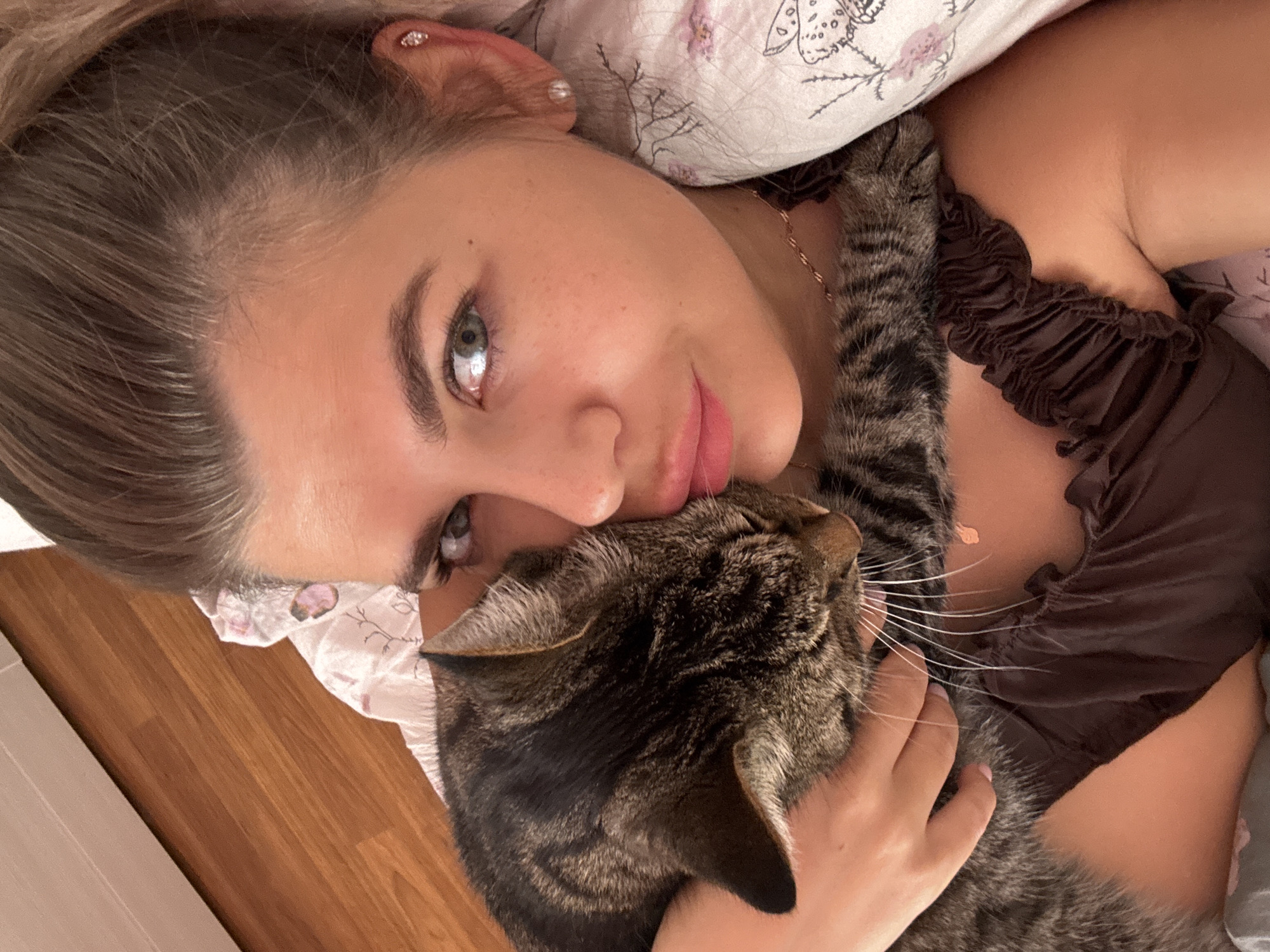 The image is a close-up selfie of a young woman with a tabby cat. The woman is lying down, holding the cat close to her face. Her hair is pulled back, and she is wearing a brown top. The cat is nuzzling her cheek. They are on a bed with floral patterned sheets. The background shows a wooden floor and a wall, suggesting an indoor domestic setting, likely a bedroom. There is no explicit indication of the location being Chaikovsky, Russia.