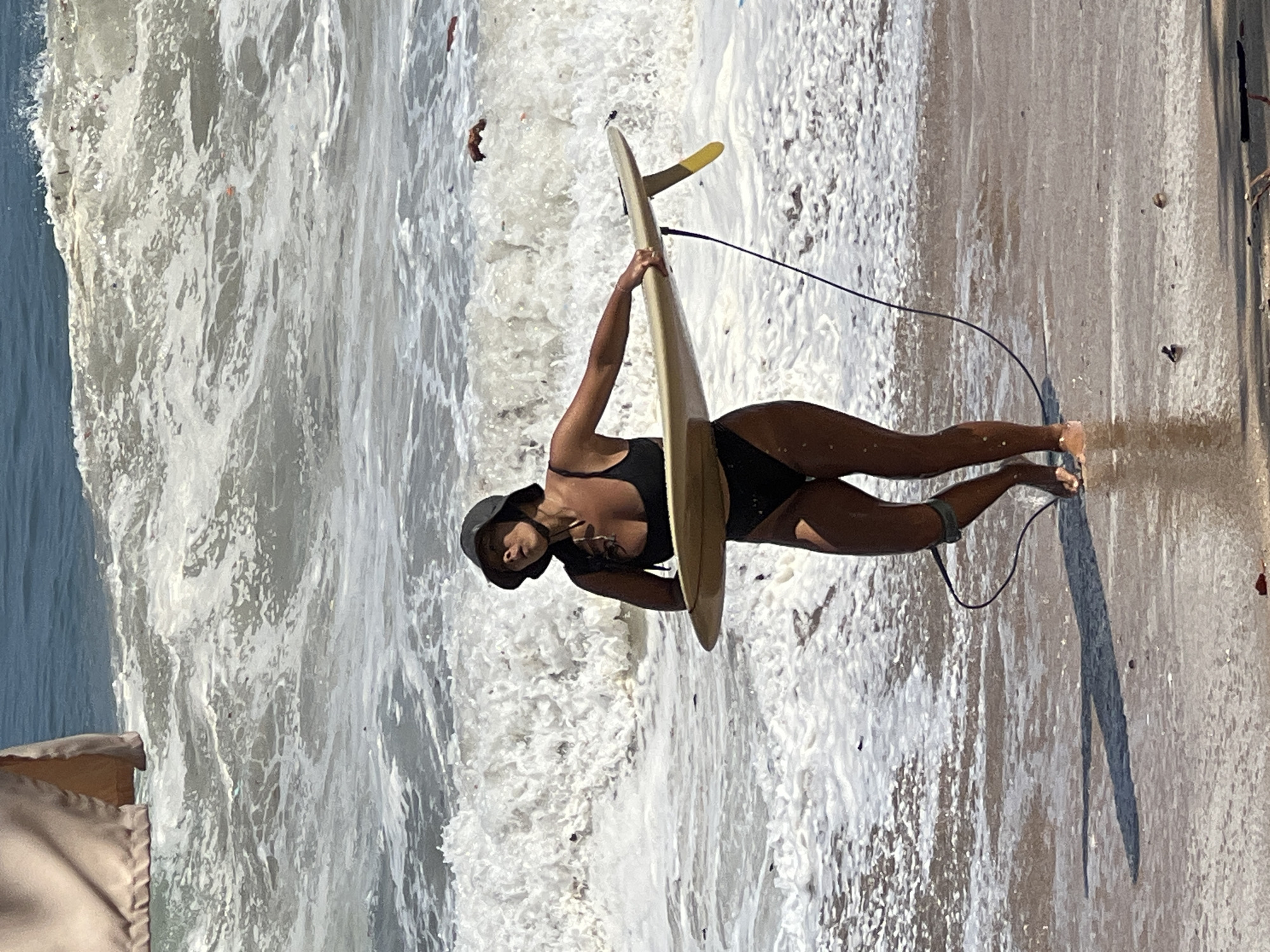 A woman in a black bikini and a dark bucket hat stands on a sandy beach in Kuta, Indonesia, holding a light-colored surfboard with a yellow fin. She is facing left, with the surfboard angled away from her body, its tail pointing to the right. The woman's eyes are closed, and her head is tilted back slightly. She has a dark leash attached to her ankle, which connects to the surfboard. The ocean behind her is rough, with large white waves crashing and foamy water reaching the shore. The sand in front of her is wet from the incoming tide, with reflections of the light on the surface. A portion of a tan fabric structure is visible in the upper left corner of the frame.