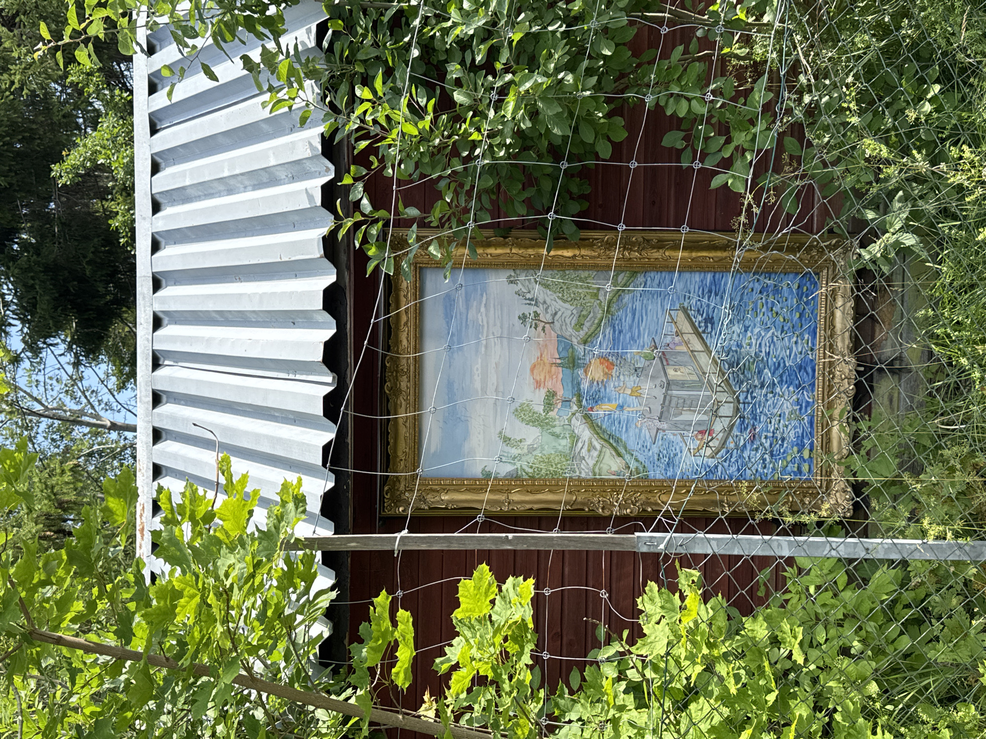 An outdoor painting is mounted on the side of a dark red wooden building, partially obscured by green foliage and a wire fence. The painting, encased in an ornate gold frame, depicts a blue body of water with a boat and figures, set against a backdrop of hills and a sunset sky. The building has a corrugated metal roof. The scene suggests a rustic outdoor setting, possibly a garden or yard in Åkersberga, Sweden.