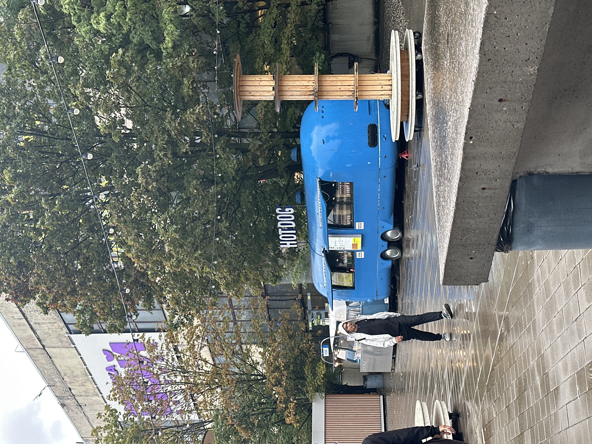 In Saint Petersburg, Russia, on a wet, overcast day, a food truck scene unfolds in an urban outdoor space. A bright blue, retro-style food truck, adorned with a "HOT DOG" sign, is prominently positioned, its serving window open. Adjacent to it, a person wearing a light-colored hooded jacket and dark pants walks by, their face turned slightly away from the camera. Another food stand, smaller and silver, is visible to the left of the blue truck. Behind the food trucks, trees with lush green foliage and some autumnal colors suggest the season. In the background, a large, utilitarian building with a weathered facade and purple lettering stands tall. The ground is paved with square tiles that glisten with moisture, reflecting the overcast sky and the surrounding structures, indicating recent rain. Decorative string lights are suspended between trees and buildings, adding a festive element to the otherwise subdued atmosphere. A tall, wooden spool structure, serving as a display or seating area, stands to the right of the hot dog truck. The overall scene depicts a casual outdoor dining area in an urban environment, likely a public square or market, with a focus on food vendors and pedestrian activity.