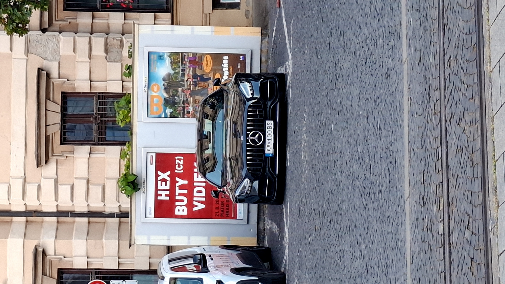 A black Mercedes-Benz AMG GT sedan is parked on a cobblestone street in front of a building with classical architecture. The car has a license plate that reads "AA 100 BS". Behind the car are two large advertisements. The advertisement on the left is red with white text that says "HEX BUTY (CZ) VIDIE". The advertisement on the right features an illustration of people and elephants. A white van is parked to the left of the Mercedes. The image was taken in Bratislava, Slovakia.