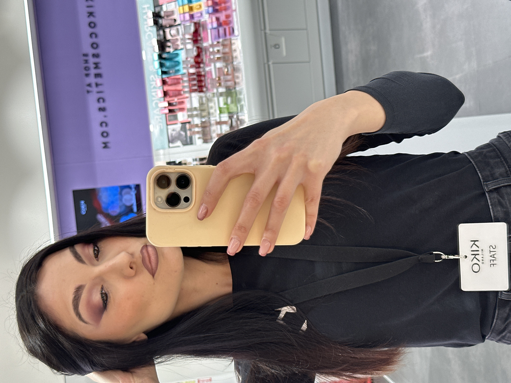 A woman is captured in a selfie taken in a mirror, likely inside a KIKO Cosmetics store. She has long, dark hair, dark eyebrows, and is wearing prominent makeup, including purple eyeshadow and nude-toned lipstick. Her nails are manicured with a delicate light pink gradient. She wears a black long-sleeved top and dark jeans.
She is holding a light yellow smartphone with a triple camera array, taking the photo. Around her neck hangs a lanyard with a clear "KIKO STAFF" ID badge. The badge clearly shows the "KIKO" logo, with "MILANO" beneath it, and the word "STAFF" (appearing mirrored as "ЭFАТS" in the reflection).
The background, visible through the mirror's reflection, reveals shelves stocked with various colorful cosmetic products, typical of a beauty retail environment. A prominent purple sign is also reflected, displaying the text "SHOP YA" and "KIKOCOSMEITCS.COM," further confirming the location as a KIKO Cosmetics establishment. The setting is indoors, well-lit, and appears to be a professional retail space.