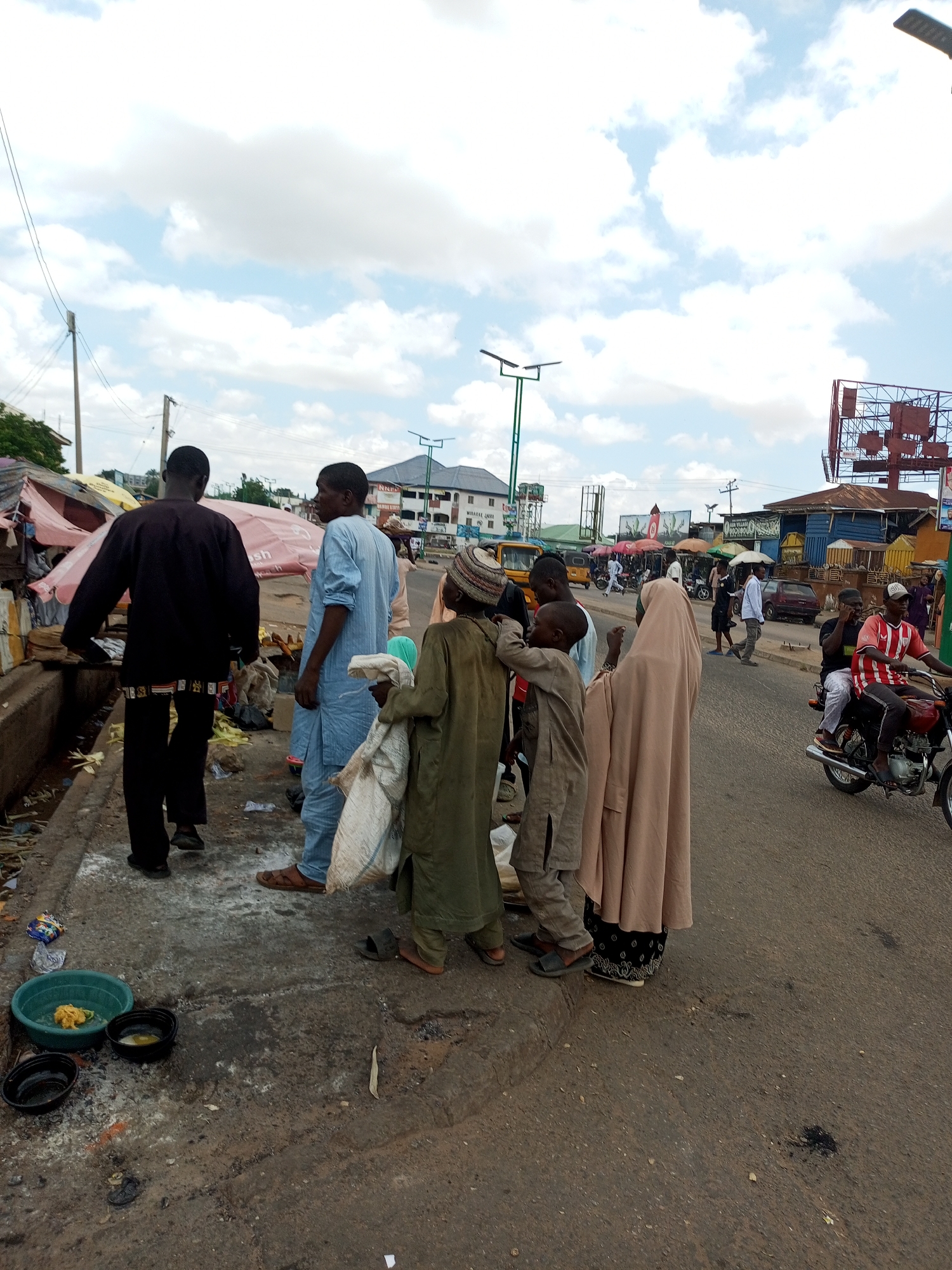 The image depicts a street scene in Arawa, Nigeria. Several individuals, predominantly men and boys, are gathered on the side of a paved road. In the foreground, a man in a black outfit stands with his back to the camera, facing away from the road. Beside him, a man in a light blue tunic and trousers is looking towards the right. A group of younger individuals are clustered near him, one of whom is carrying a large white sack. Another boy, wearing a light brown garment and a cap, stands to the right, with a portion of his face visible. A woman in a light brown hijab and a patterned skirt is also present.
The environment appears to be a busy marketplace or roadside area, with market stalls and awnings visible in the background. Debris and refuse are scattered along the edge of the road and pavement. In the middle ground, various vehicles, including a yellow tricycle and a motorcycle, are visible. The sky is partly cloudy. The overall impression is of an active, albeit somewhat disorganized, urban environment.