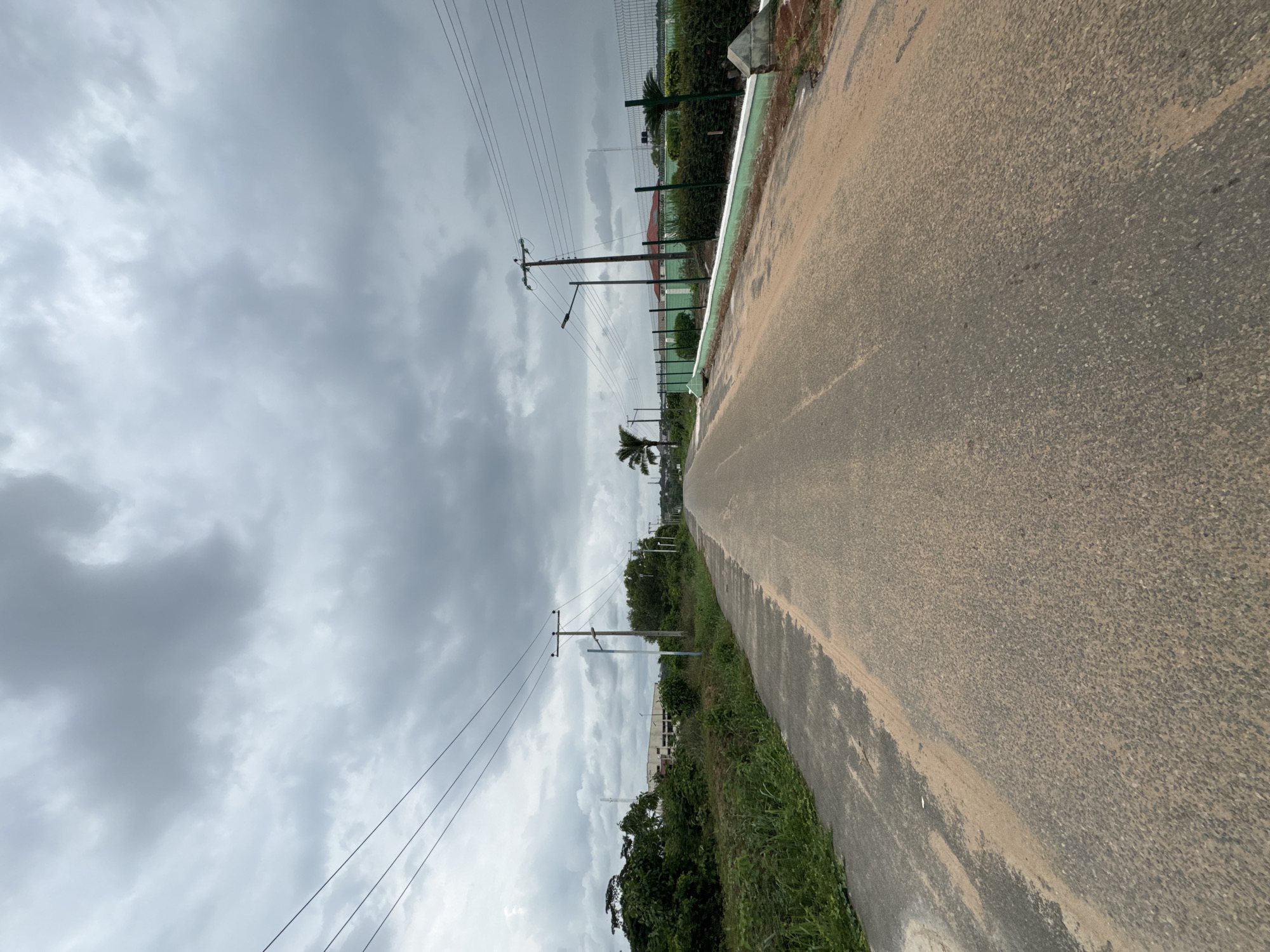 The image depicts a straight, paved road in Ihiagwa, Nigeria. The road surface appears to be asphalt, with a noticeable accumulation of sandy material along the edges. Overhead, power lines stretch along the road's length, supported by wooden utility poles. The sky is heavily overcast with gray clouds. Vegetation, including grassy areas and shrubs, lines the sides of the road. In the distance, fencing and some low-rise buildings are visible. There are no people, animals, or vehicles present in the scene.