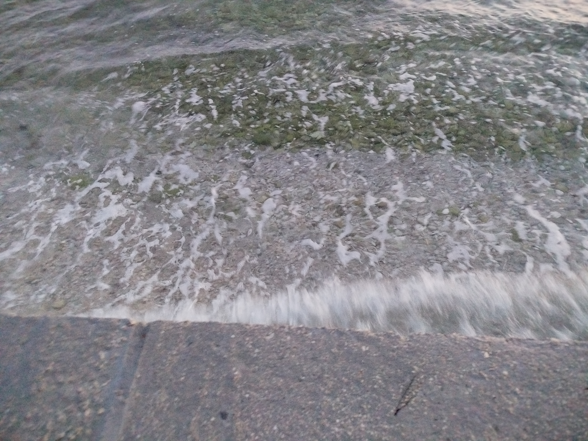 The image captures a close-up, slightly blurred view of the sea meeting a paved area. The water is shallow, revealing a seabed covered in small, grey and green stones. White foam from the small waves washes over the stones and then recedes, creating a dynamic, foamy edge against the dark grey, textured paving. The paving appears to be made of concrete or stone slabs, with a distinct edge separating it from the water. A faint shadow is cast on the paving. The overall lighting suggests it is either early morning or late evening. The scene is likely a waterfront promenade or quay in the city of Brist, Croatia, characterized by its pebbly seabed and accessible paved edge. There are no people or animals visible in the image.