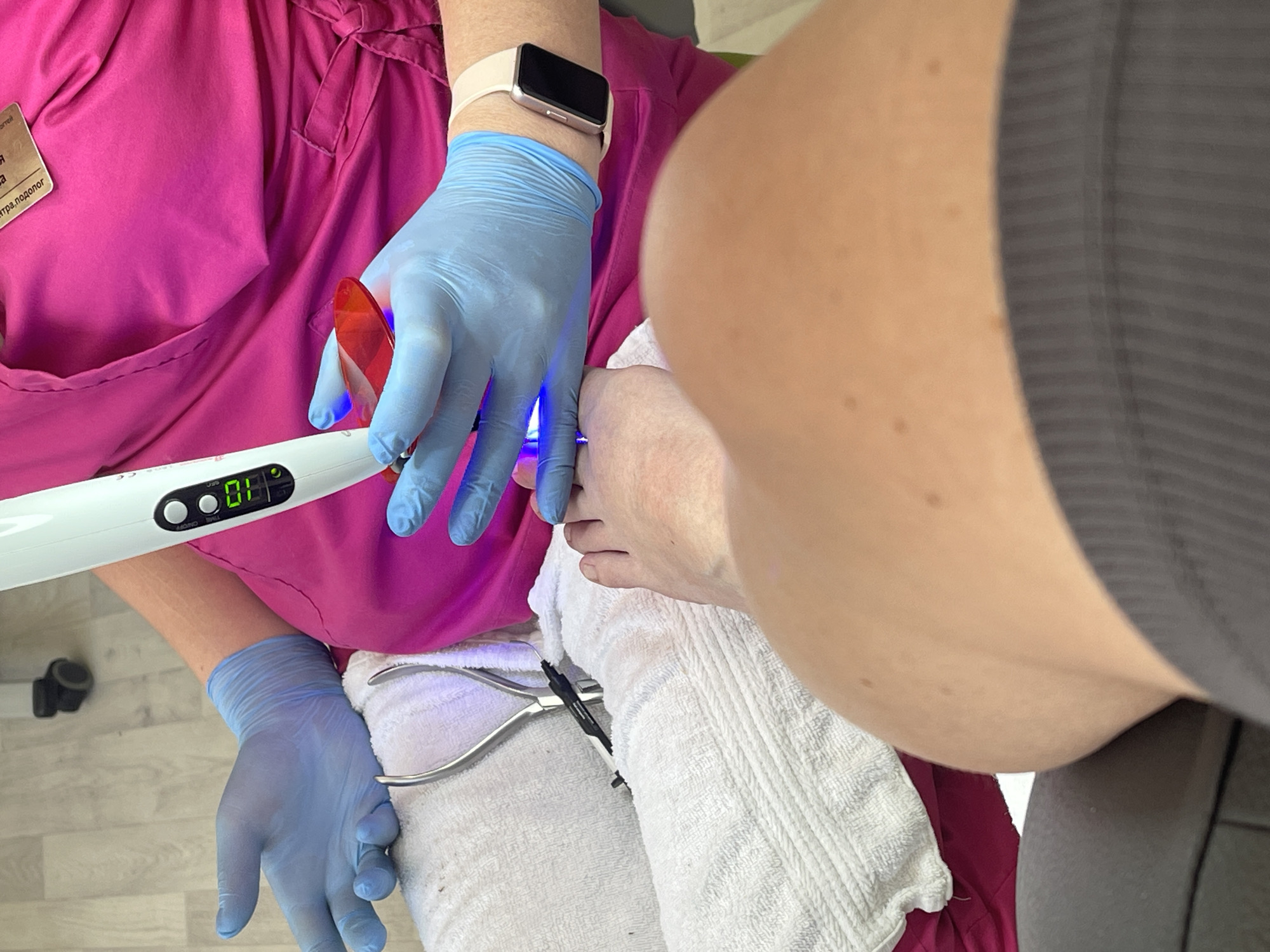 In an indoor setting, an individual wearing blue medical gloves is using a UV light device on a person's foot. The UV light device has a digital display showing "08.8". The individual is dressed in a bright pink scrub top, and a white towel is placed under the foot. A pair of metal nail nippers are visible on the towel. The perspective is from the person receiving the treatment, looking up towards the practitioner and their own foot. The environment appears to be a salon or clinic.