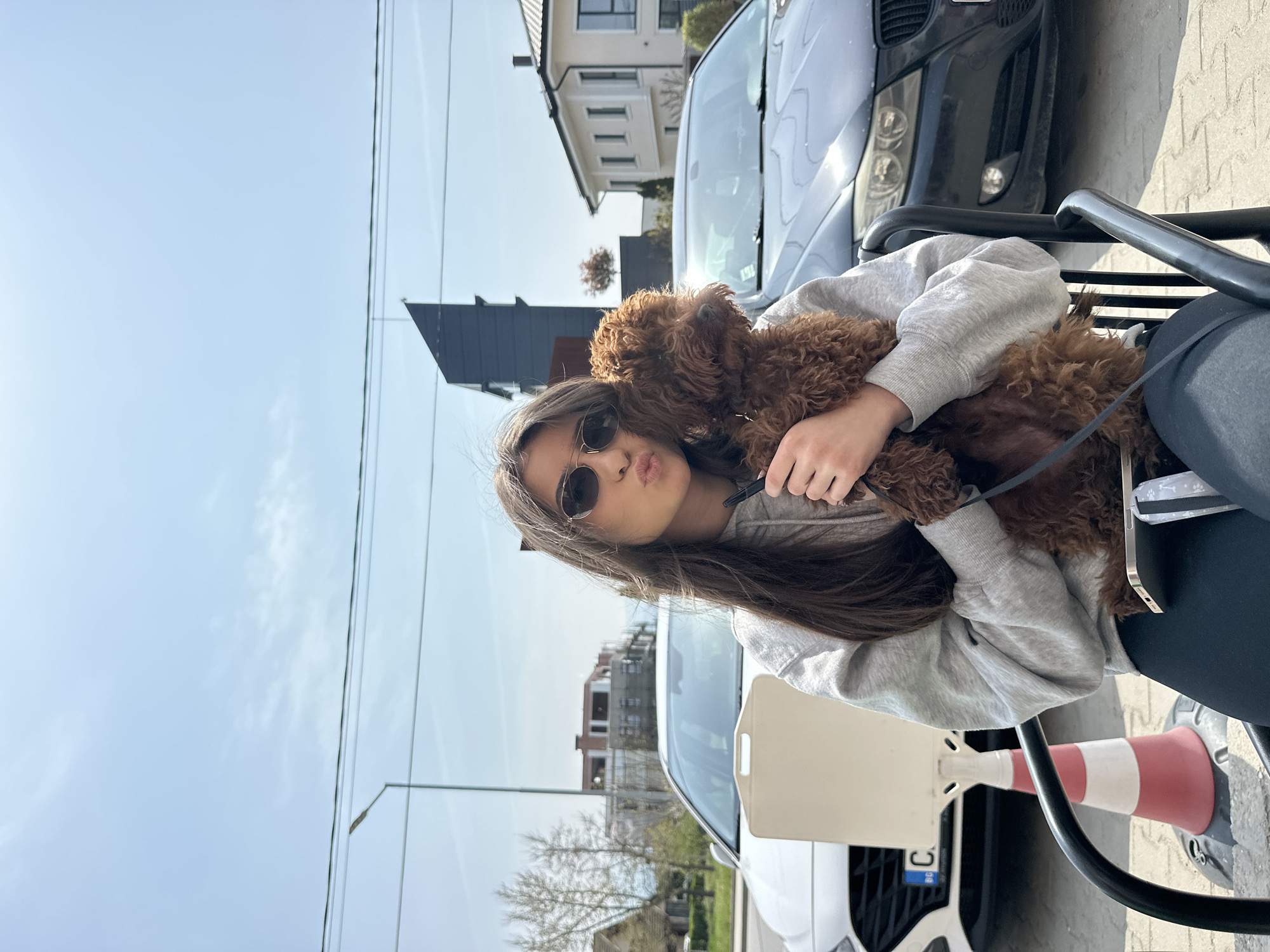 A young woman is sitting on a metal chair and holding a fluffy brown puppy. She is wearing a light gray hooded sweatshirt, dark leggings, and sunglasses. She is making a kissing face towards the camera. A leash is attached to the dog's collar. Next to the chair is a white and red traffic cone. Two cars are partially visible behind her: a dark-colored car and a white car. The background shows buildings and a blue sky. The photograph was taken outdoors in what appears to be an urban area.
The location is Plovdiv, Bulgaria.