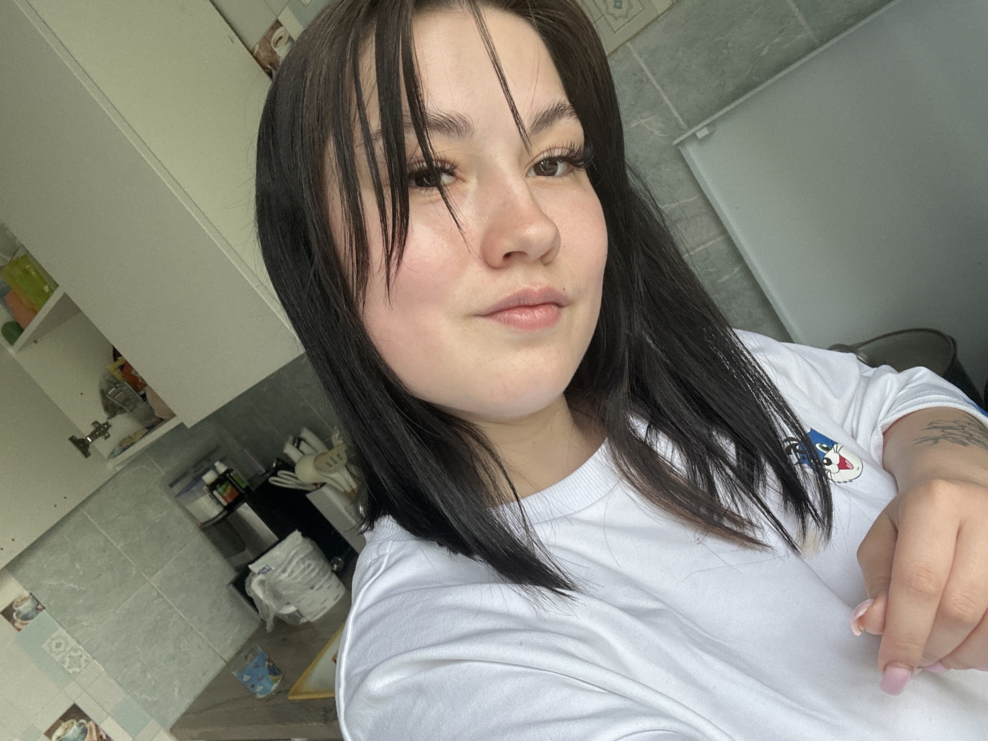 The image shows a young woman with dark hair, a fair complexion, and neutral lipstick, posing for a selfie. She wears a white T-shirt with a cartoon graphic. The background reveals a domestic kitchen setting with white cabinetry and tiled walls. Kitchen utensils and items are visible on the countertop. Based on available information, the location of the image is Chaikovsky, Russia.
