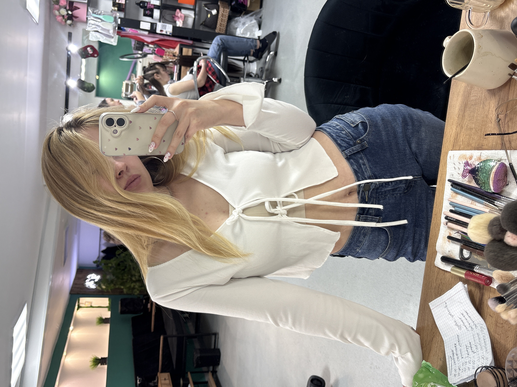 A woman is taking a selfie in what appears to be a salon. The woman has long blonde hair, and she is wearing a white top and blue jeans. She is holding a phone with a heart-patterned case, partially obscuring her face.
In the background, other people can be seen sitting in chairs and getting their hair done. There are mirrors, styling tools, and salon equipment visible. A notebook with writing is visible on a table, along with makeup brushes and other beauty products.
The photo was taken in Chernushka, Russia.