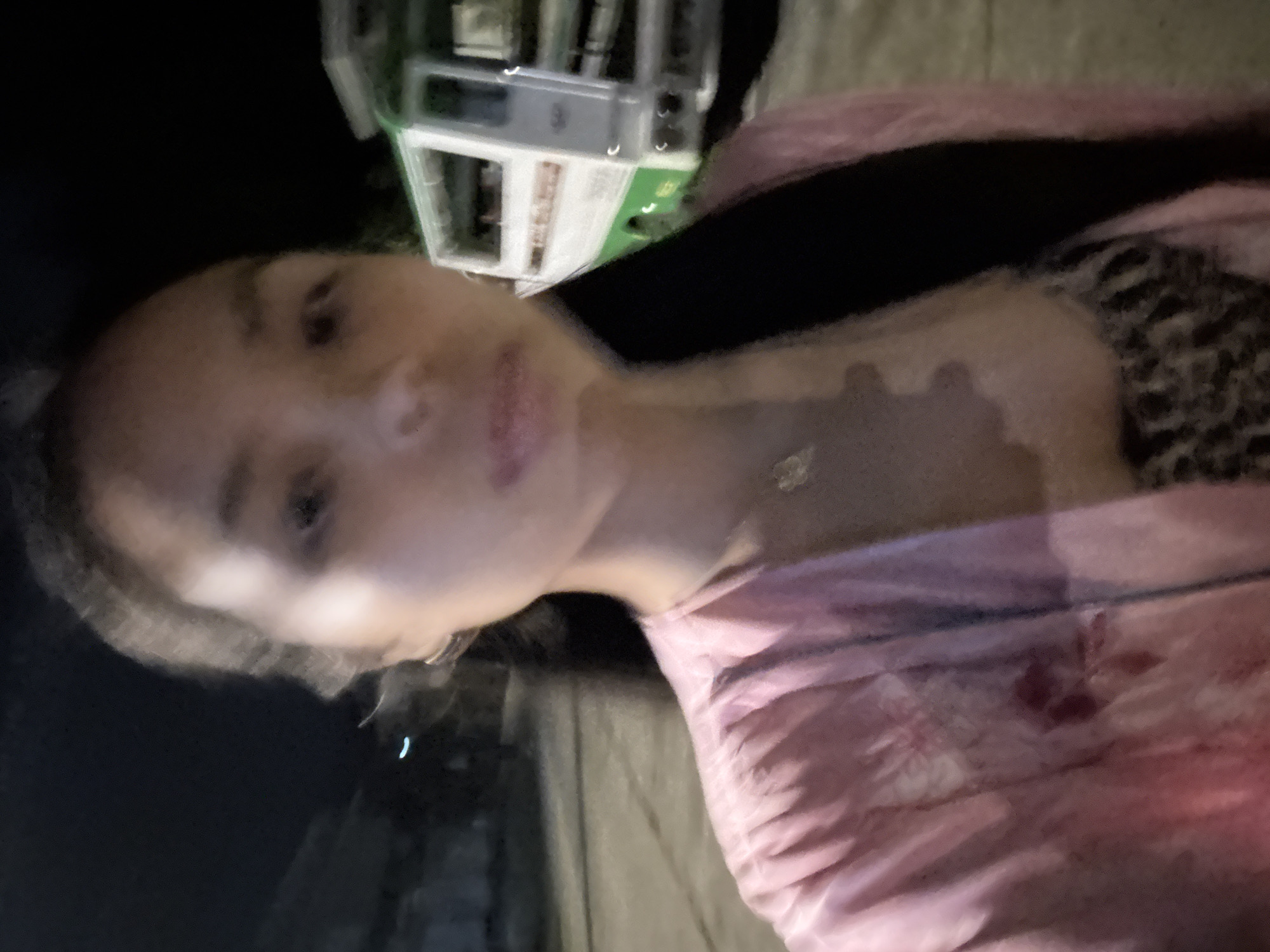 A blurry, low-light image depicts a person with fair skin and dark hair looking towards the camera. They are wearing a pink garment with what appears to be a black strap over their shoulder. A building with green and white elements is visible in the upper left corner. The setting appears to be outdoors, potentially a street or sidewalk at night. The city and country were provided.