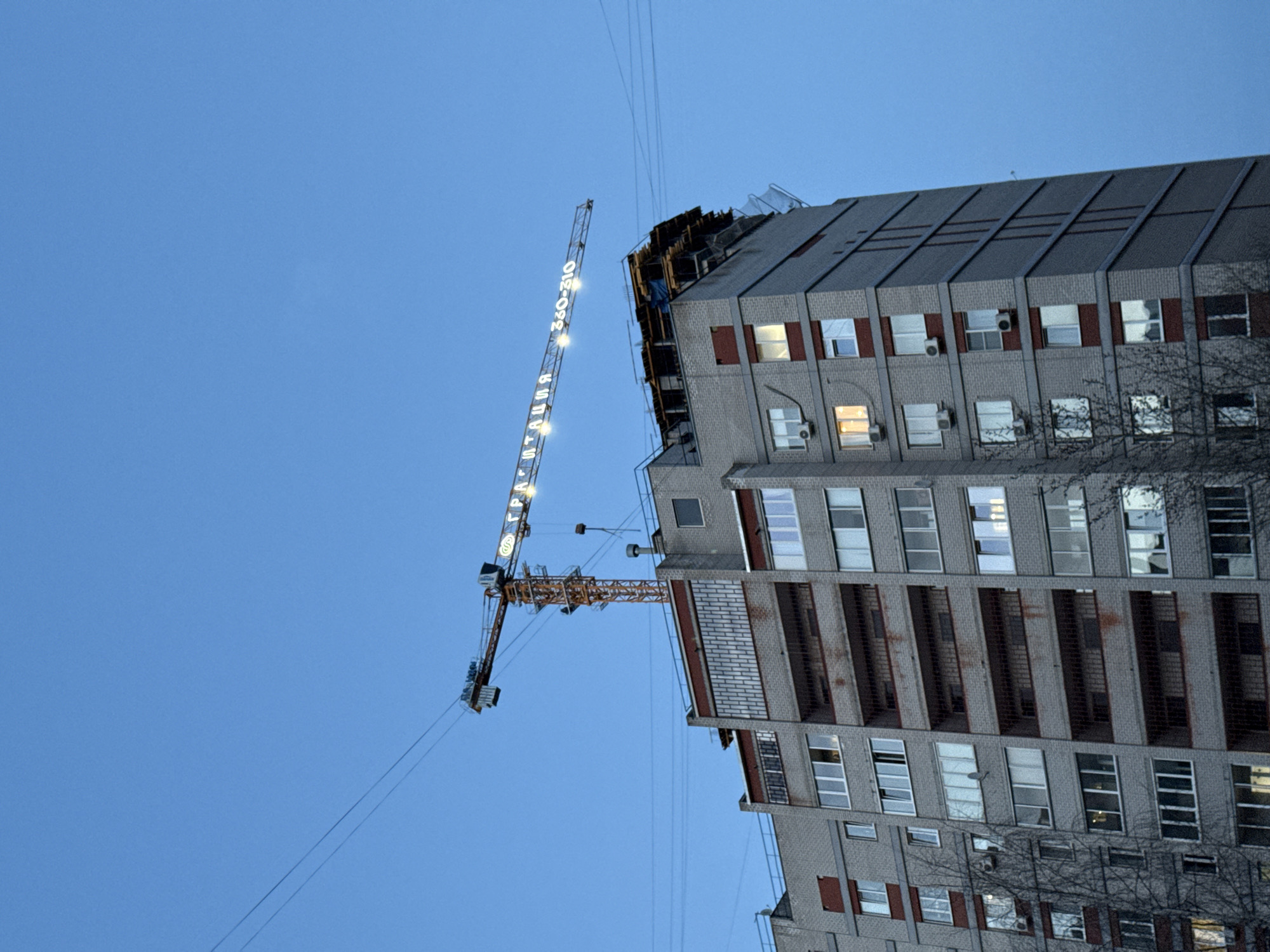 The image shows a tall building under construction in Izhevsk, Russia. A construction crane is positioned next to the building. The building has a grey brick facade with rows of windows. Some windows have red trim. Several air conditioning units are mounted on the exterior walls. The sky is a light blue, suggesting it is either early morning or evening. A tree is partially visible at the bottom right corner. The crane is lit up with lights.