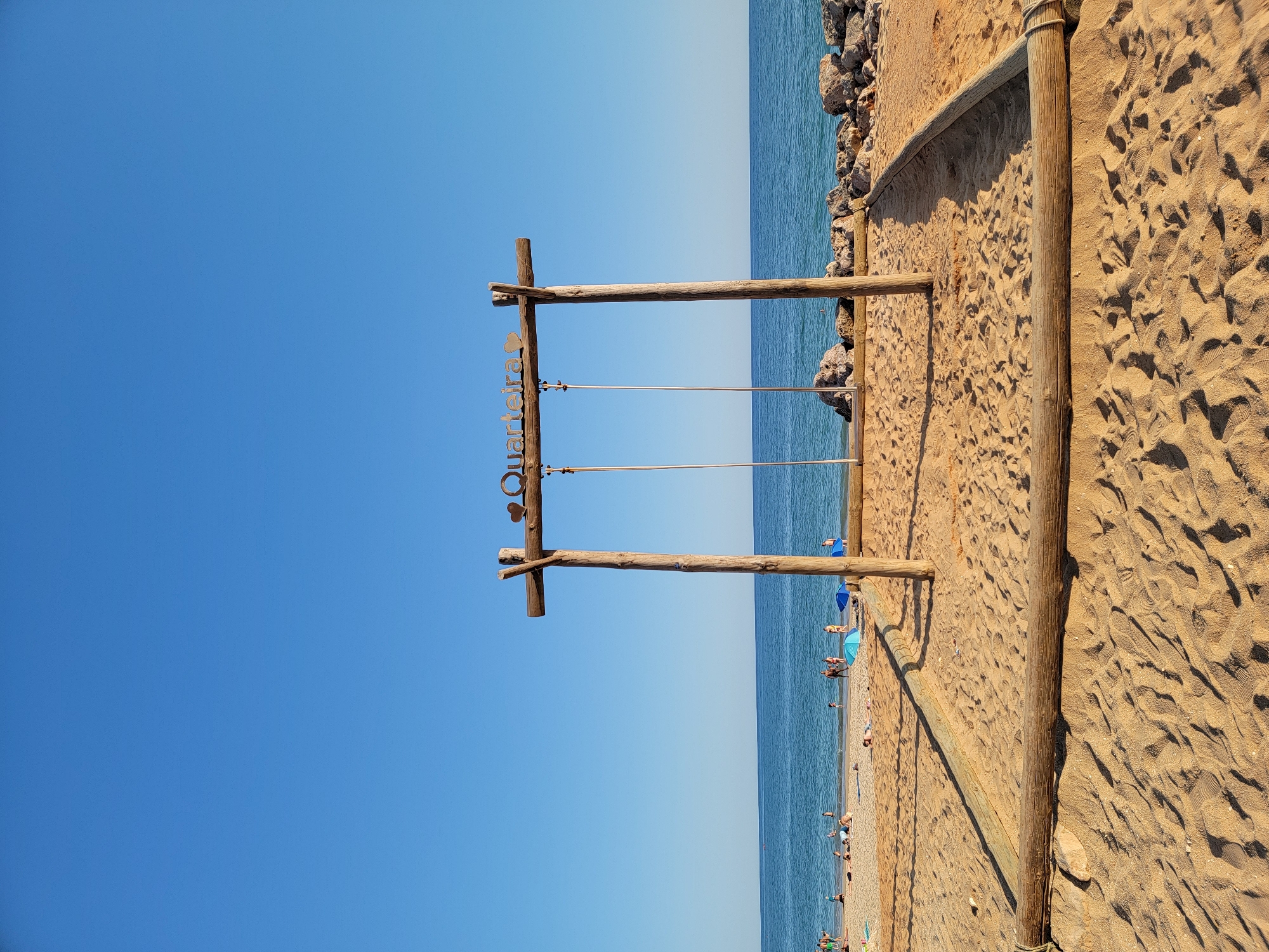 The image depicts a beach scene in Quarteira, Portugal, dominated by a wooden swing set positioned centrally on the sand. The swing set features two vertical wooden posts supporting a horizontal beam, from which two sets of ropes hang, indicating the presence of swings. A sign with the text "Quarteira" and a heart symbol is affixed to the horizontal beam.
The beach is covered in golden sand, with a sandy perimeter marked by logs. In the background, the blue ocean stretches towards the horizon under a clear blue sky. Along the shoreline, several people are visible, some sunbathing on towels, others walking or wading in the shallow water. There are also blue beach umbrellas scattered on the sand. The foreground shows the texture of the sand, with footprints and shadows cast by the midday sun. Rocky formations are visible along the edge of the water to the right of the swing set. The overall atmosphere is one of a sunny day at a coastal resort.