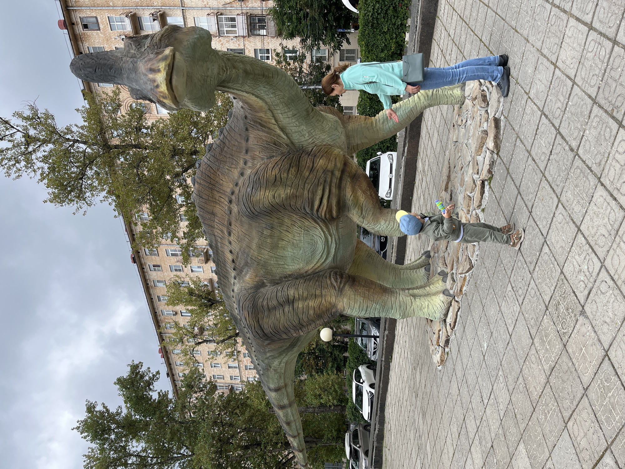 In Moscow, Russia, a life-size statue of a Parasaurolophus dinosaur stands on a paved plaza. A woman in a teal jacket and jeans and a young child in a khaki jumpsuit and blue cap are interacting with the statue. The child holds a toy. Behind the dinosaur, a tall, brick apartment building with numerous windows and balconies rises against a cloudy sky. Trees with green foliage line the street in front of the building, and several cars are parked.