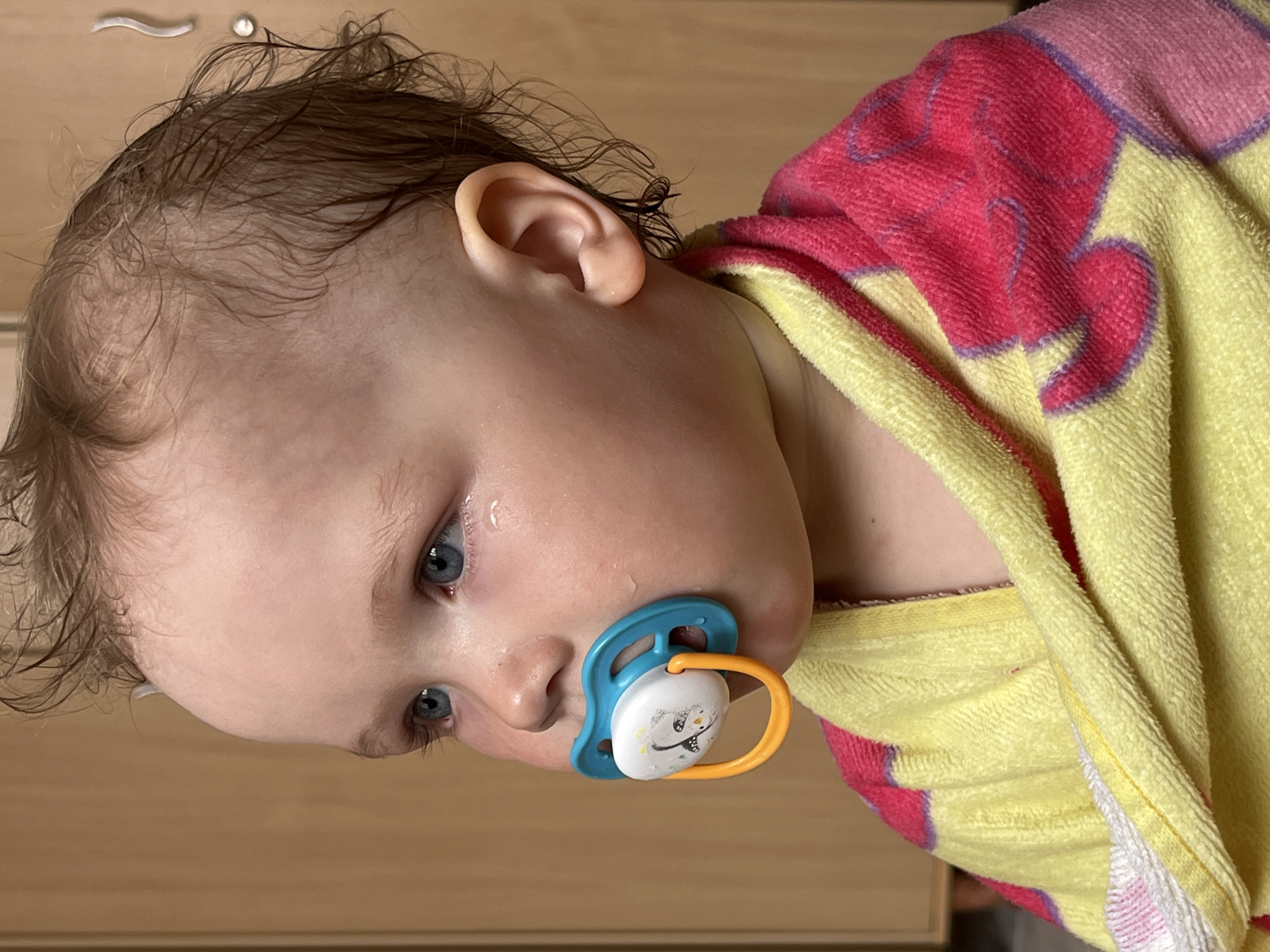 A close-up, rotated image shows a baby with light skin, blue eyes, and damp, light brown hair. The baby has a blue and yellow pacifier in its mouth. A pink and yellow patterned towel is draped around the baby's neck. The background appears to be a light brown, wood-like surface, likely a dresser or cabinet. Location: Unknown, Unknown.