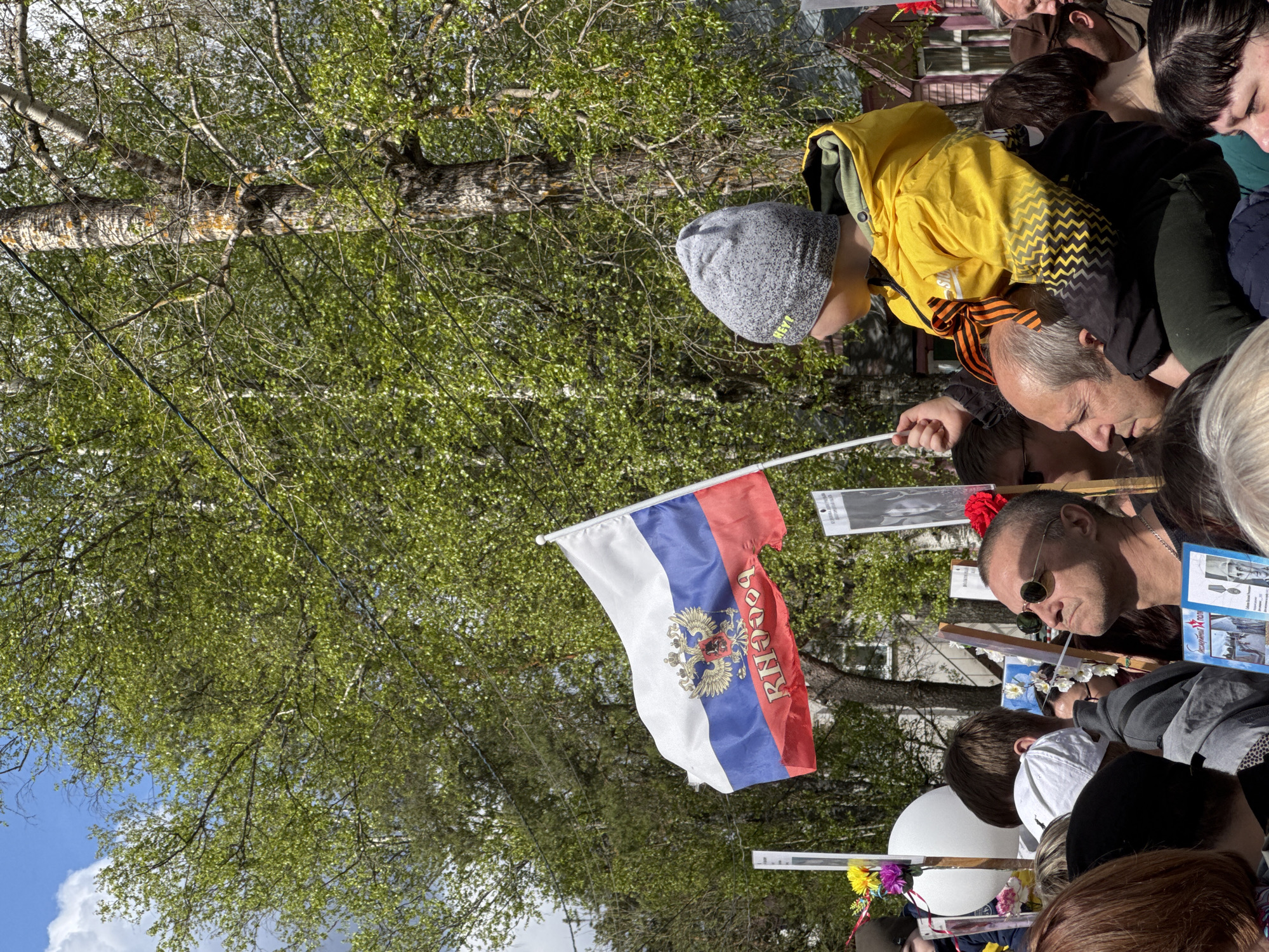 The image shows a crowd of people gathered outdoors, possibly participating in a public event or gathering in Chuprino, Russia. In the foreground, a Russian flag with the inscription "Россия" (Russia) is prominently displayed. People of varying ages are present, with some individuals holding up framed photographs or signs. A young boy in a yellow jacket and a grey hat stands near an adult who is wearing a St. George ribbon. The environment includes trees with green foliage in the background, suggesting it could be springtime. The overall scene conveys a sense of community or shared event.