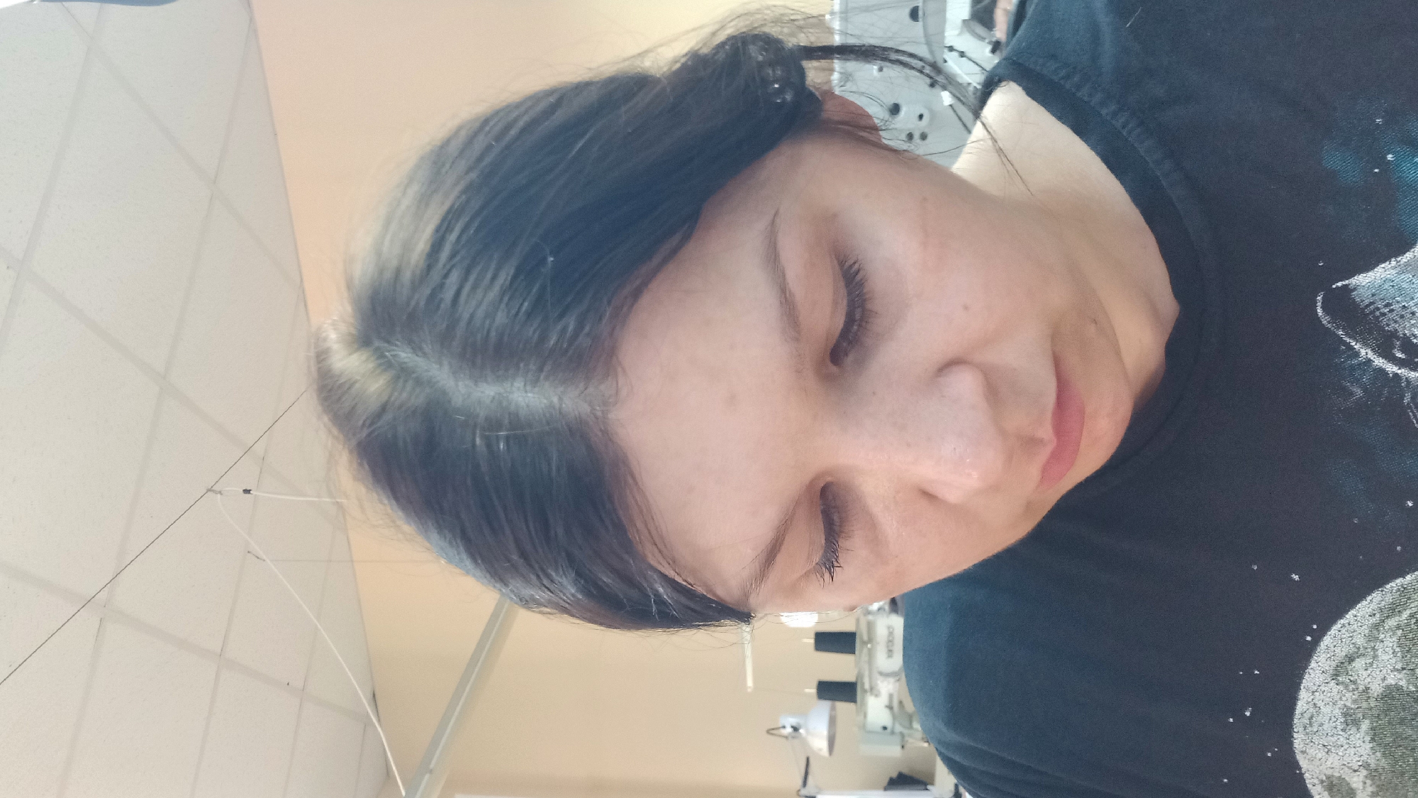 A close-up, overhead shot shows a woman looking down, her face filling most of the frame. She has dark hair parted in the middle, and her eyes are closed. She is wearing a black t-shirt with a graphic print of a moon and what appears to be a wolf. In the background, industrial sewing machines and lamps are visible, suggesting a workshop or factory setting. The ceiling is a standard drop ceiling with fluorescent lighting. There are wires hanging from the ceiling, possibly for the lighting or equipment. The overall environment appears to be indoors, likely a place of work. The location is not specified within the image.