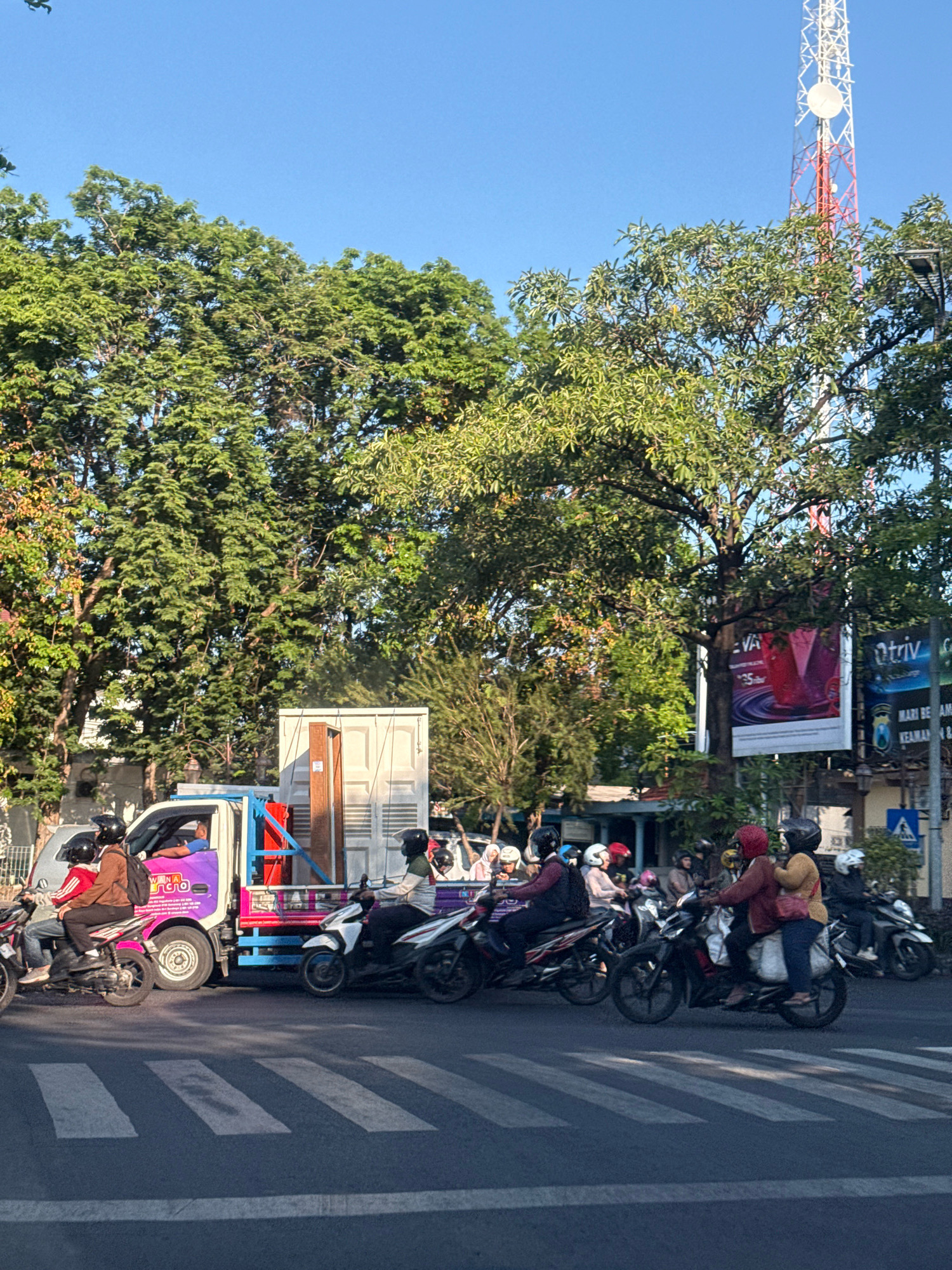 This is an eye-level, medium shot of a street scene in Surabaya, Indonesia, on a clear day with a bright blue sky. The foreground features a pedestrian crossing. A white truck with a light blue frame and a colorful advertisement on its side is stopped at the crossing, partially obscuring a group of motorcycles and their riders. The motorcycles are diverse in color, with many riders wearing helmets. Behind the truck and motorcycles, a dense line of trees with lush green foliage stretches across the middle ground. In the background, a tall, red and white telecommunications tower rises against the sky, and various buildings and advertisements are visible. The overall impression is a busy urban street with typical Indonesian traffic.