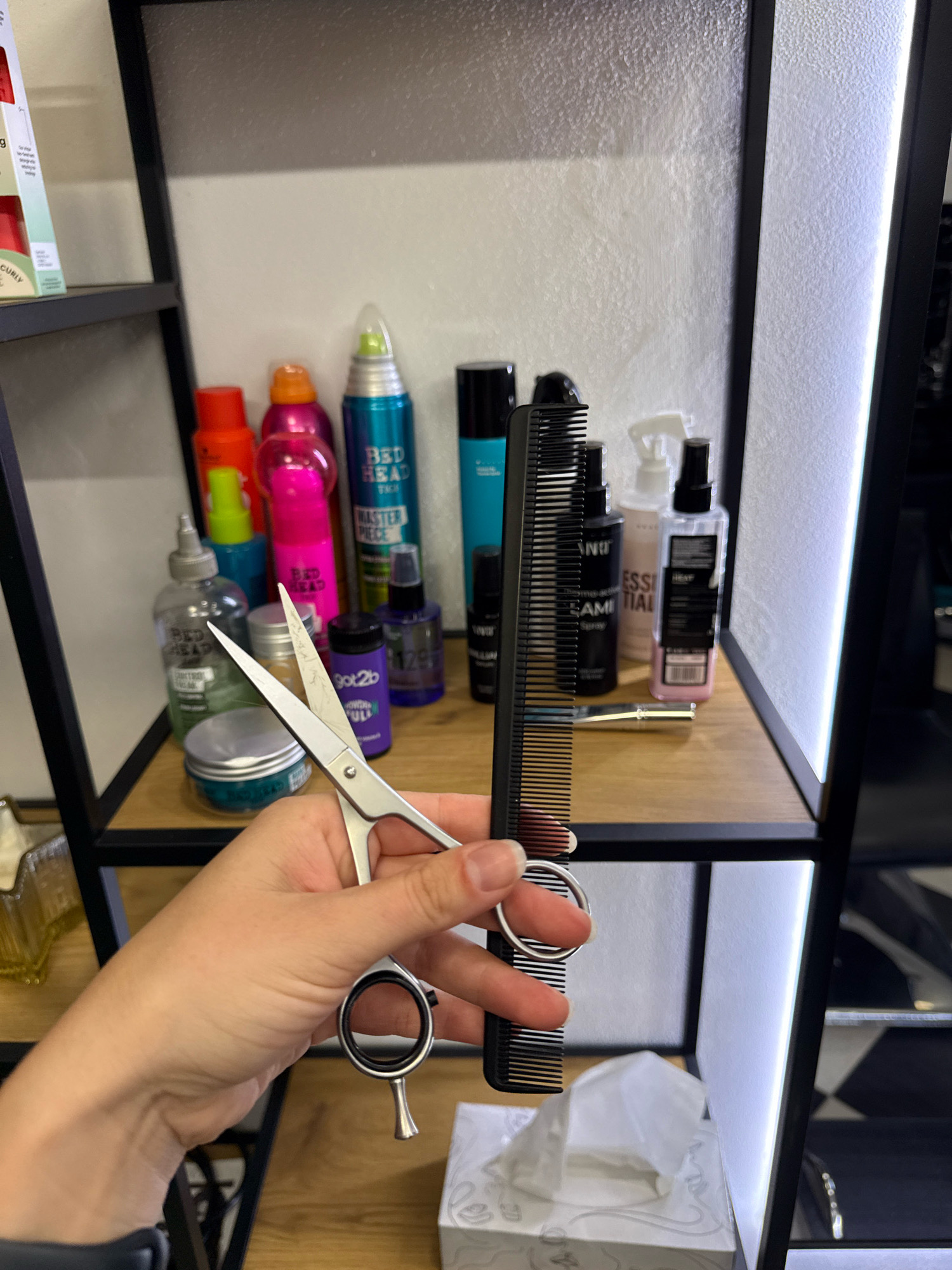 The image shows a shelf unit in what appears to be a salon or bathroom. A hand is holding a pair of silver scissors and a black comb in front of the shelves. The shelves are stocked with various hair and beauty products, including spray cans, bottles, and a jar. The products are arranged in a somewhat disorganized manner. The background consists of a white, textured wall with integrated lighting on the right side. The overall impression is one of a functional space used for hair care.