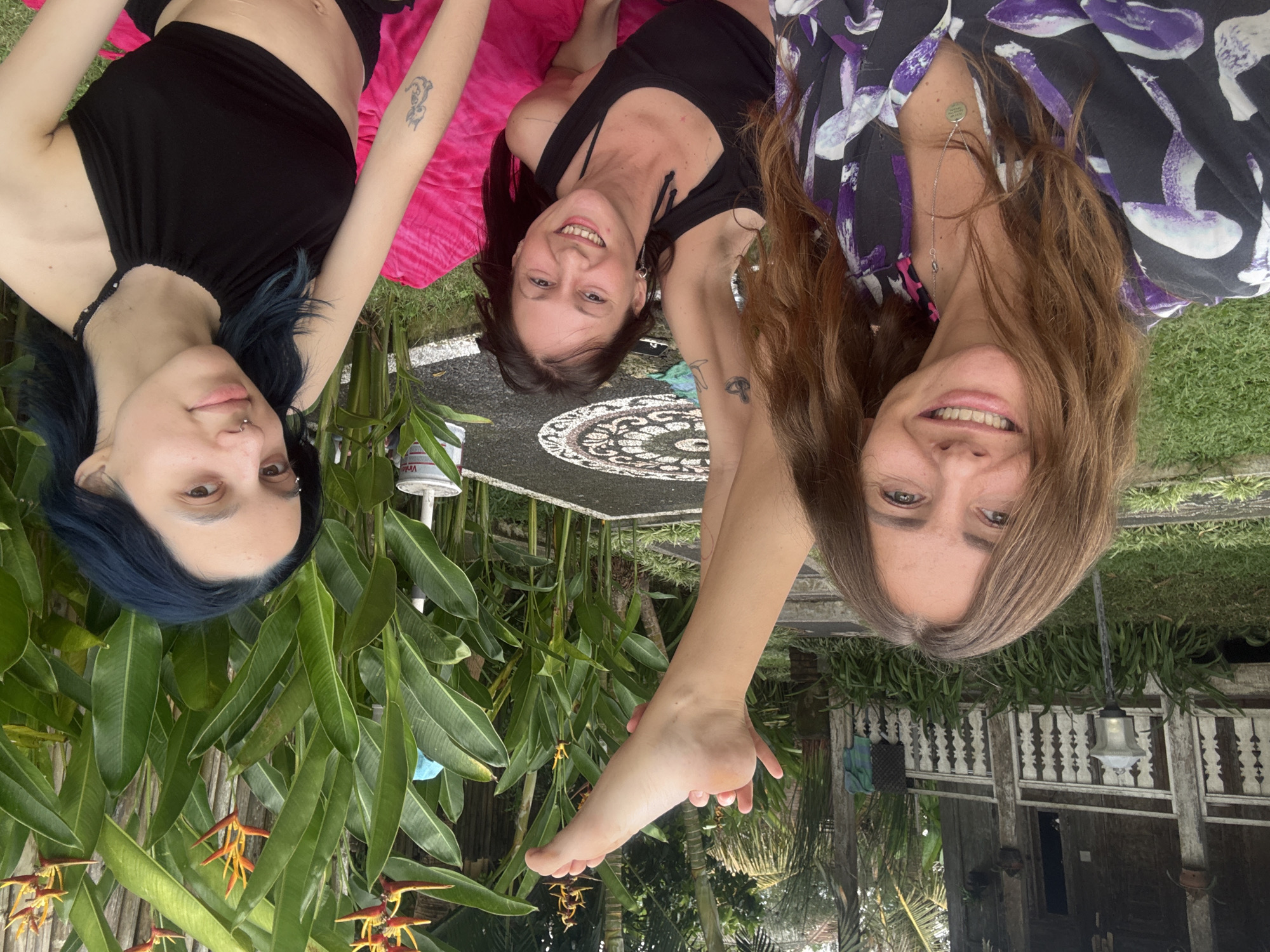 Three women are lying on the grass in Mengwi, Indonesia. The woman on the left has blue hair, wears a black top, and has a tattoo on her left arm. She lies on a pink blanket. The woman in the middle wears a black top and has a tattoo on her left arm. She lies next to a patterned table. The woman on the right wears a patterned shirt with purple and white designs and has brown hair. She has a necklace. Her bare foot is raised. The background includes greenery, plants, and a building with a white railing.
