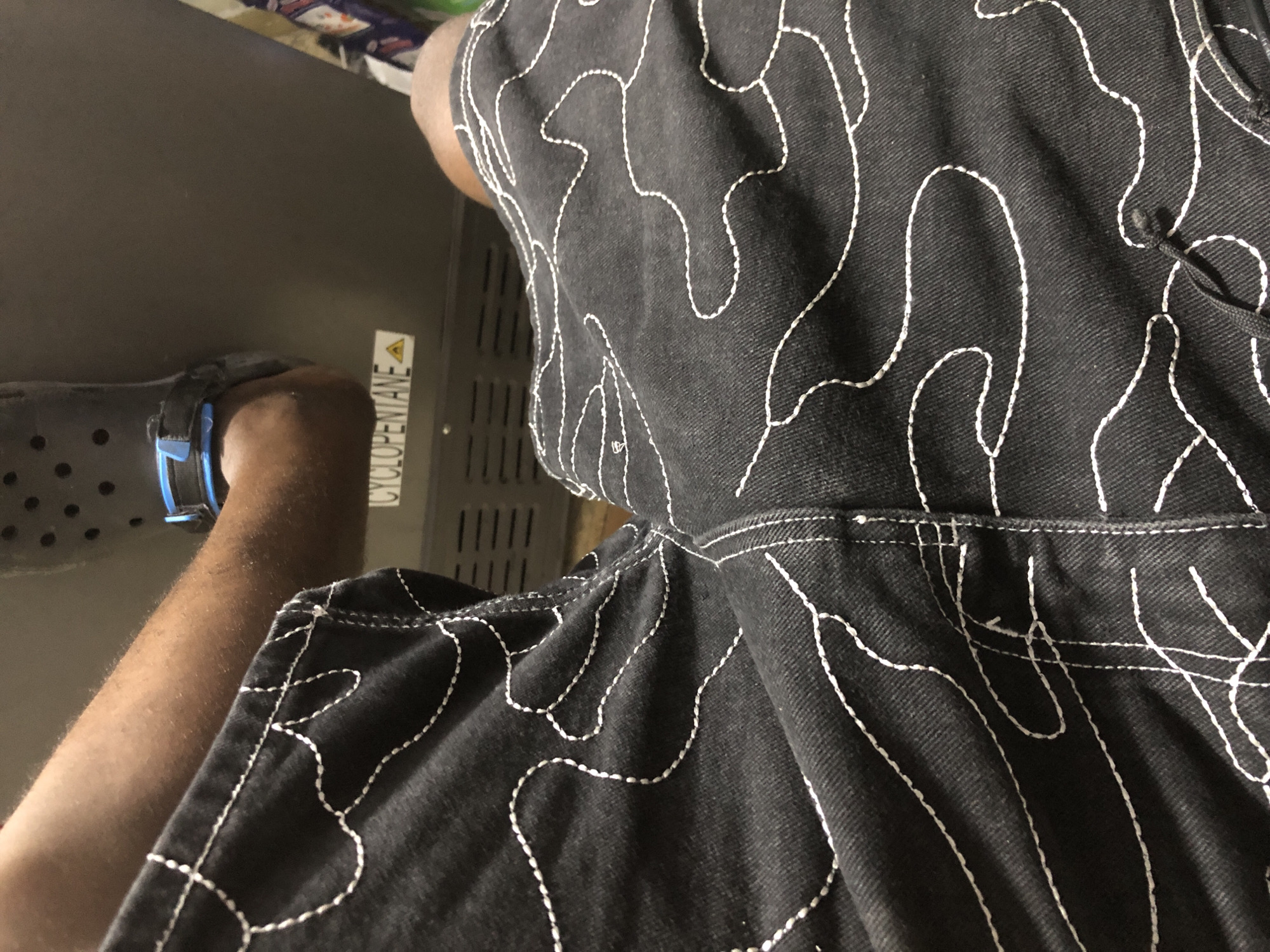 A person's legs are shown, clad in dark denim shorts with a white wavy stitch pattern. The person is wearing dark clogs on their feet. The background appears to be an interior space, possibly a bus or train, with a metal surface bearing the label "CYCLOPENTANE" and a caution symbol.
