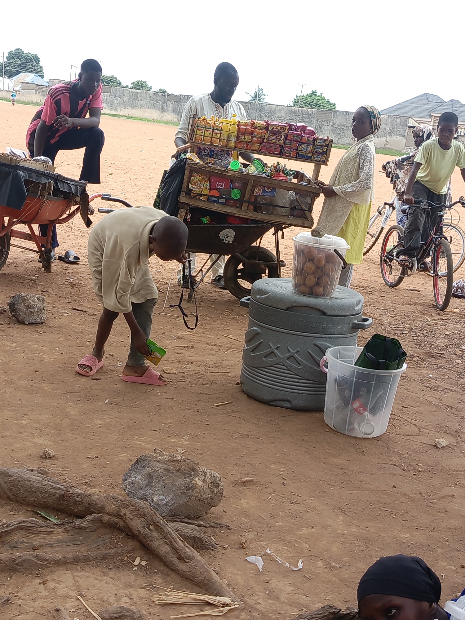 The image appears to be taken in an outdoor area in Akwanga, Nigeria, with a dirt ground surface. Several individuals are present, with a man and a young girl seemingly operating a makeshift vending cart stocked with snacks and drinks. Another man is seated on a wheelbarrow nearby. A boy in pink sandals is observed picking up an item from the ground. Another boy is riding a bicycle, with a young girl standing nearby. Visible objects include a large cooler, a bucket containing items, rocks, and tree roots. The background features buildings and a fence, indicating an urban or semi-urban setting. The scene suggests a local commercial activity or informal market.