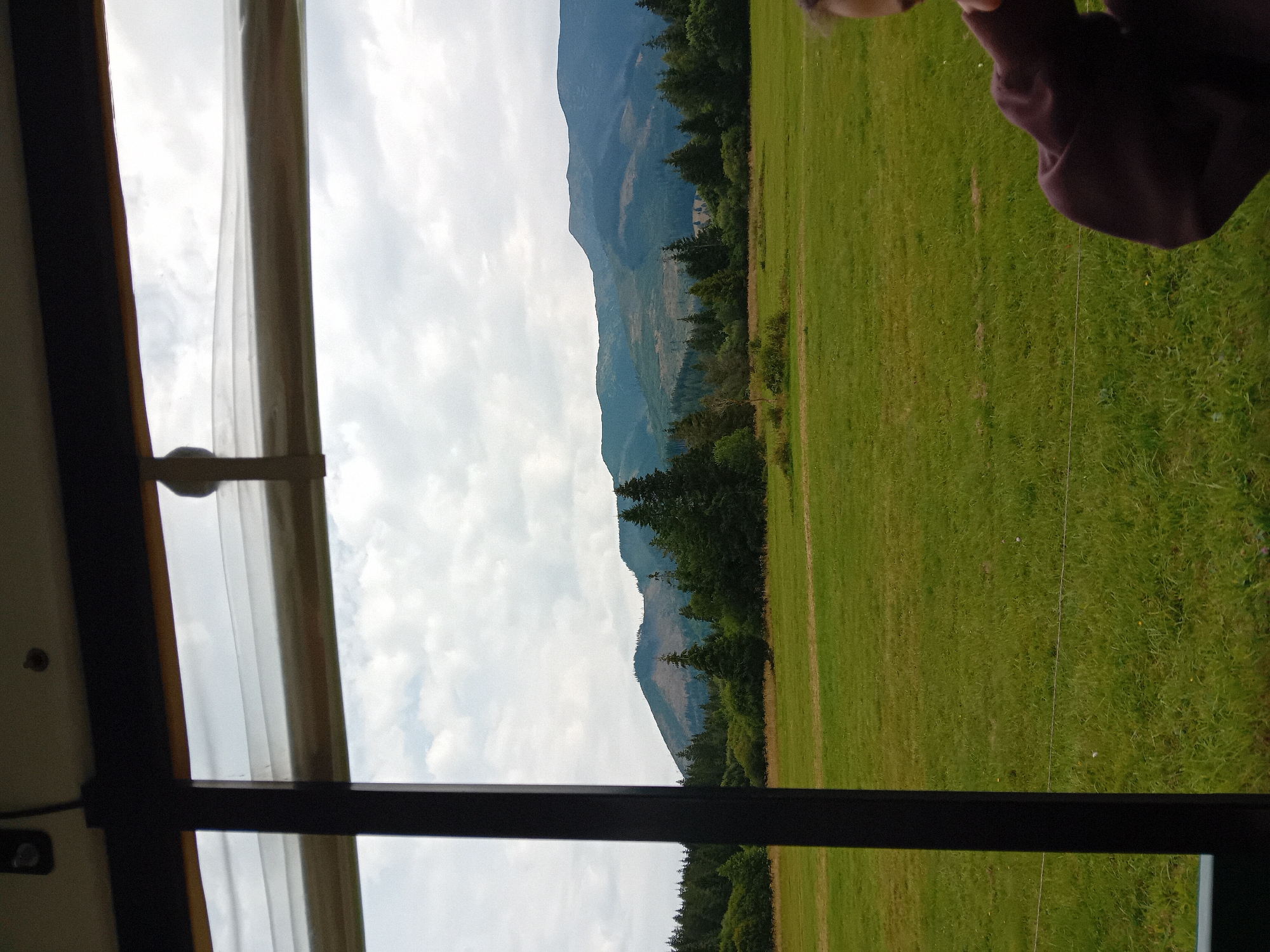 The image shows a view of mountains and a grassy field from within what appears to be a vehicle. A dark frame, possibly a window or doorway, is visible in the foreground on the left. The sky is overcast with clouds. In the distance, a range of mountains is visible, partially covered with trees. In the lower right corner of the image, a person's shoulder and head are visible, wearing a dark purple garment. The location is identified as Zuberec, Slovakia, a village in the Orava region, known for its proximity to the Tatra Mountains.