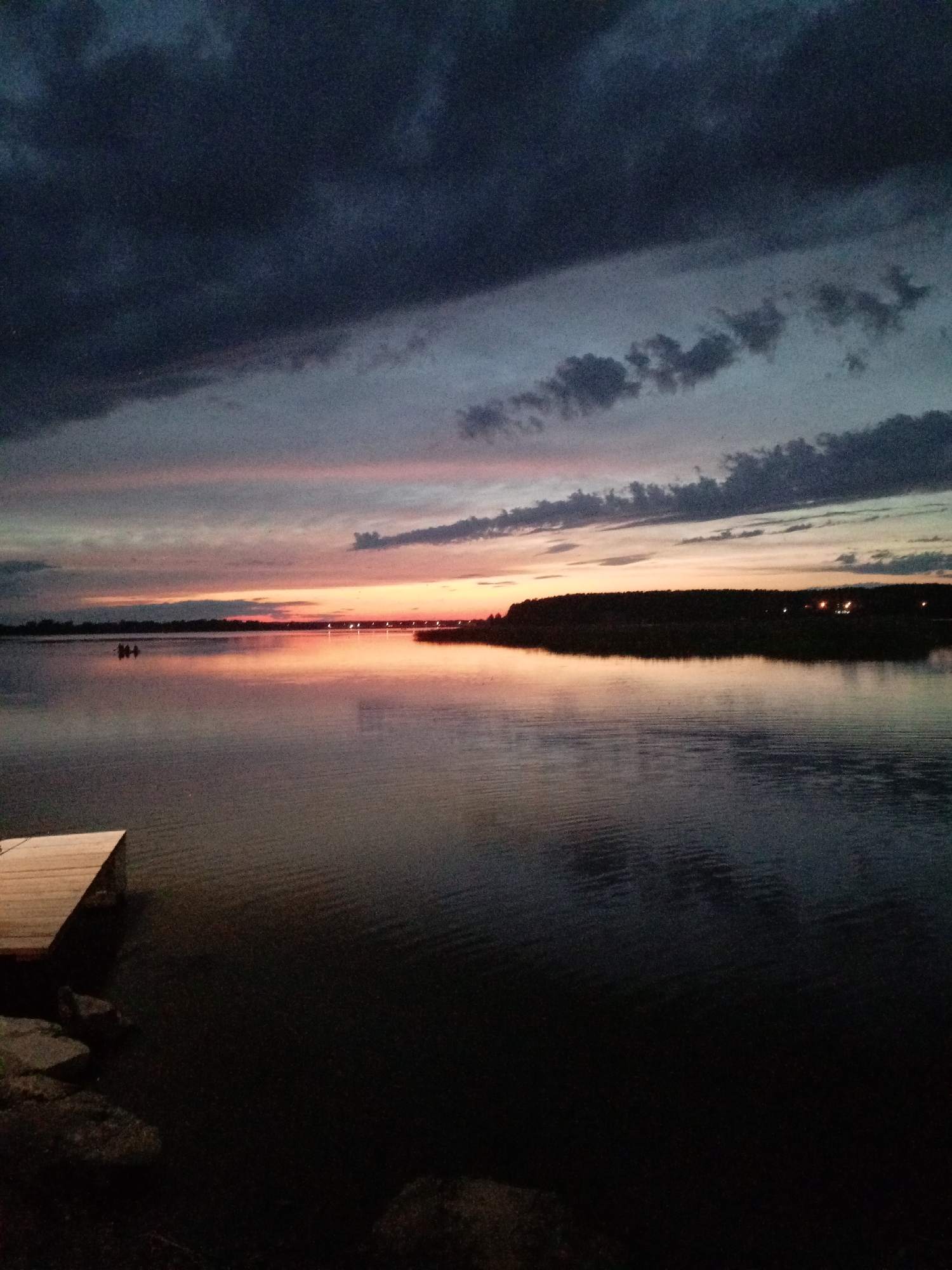 The image depicts a serene twilight scene by a body of water, likely a lake or river, with a wooden dock in the foreground. The sky is filled with dramatic clouds in shades of dark gray and deep blue, transitioning to soft pinks and oranges near the horizon, indicating sunset or sunrise. A silhouetted treeline borders the far bank of the water, with a few distant lights suggesting human habitation or structures. The water's surface is calm, reflecting the colors of the sky and creating subtle ripples. In the mid-distance, on the water, a small group of people are visible, possibly in a boat, adding a sense of scale and activity to the otherwise still landscape. The overall mood is peaceful and tranquil. This scene could be located on the shore of Lake Taimyr in Zheleznogorsk, Russia, given the natural elements present and the possibility of a tranquil waterfront environment.