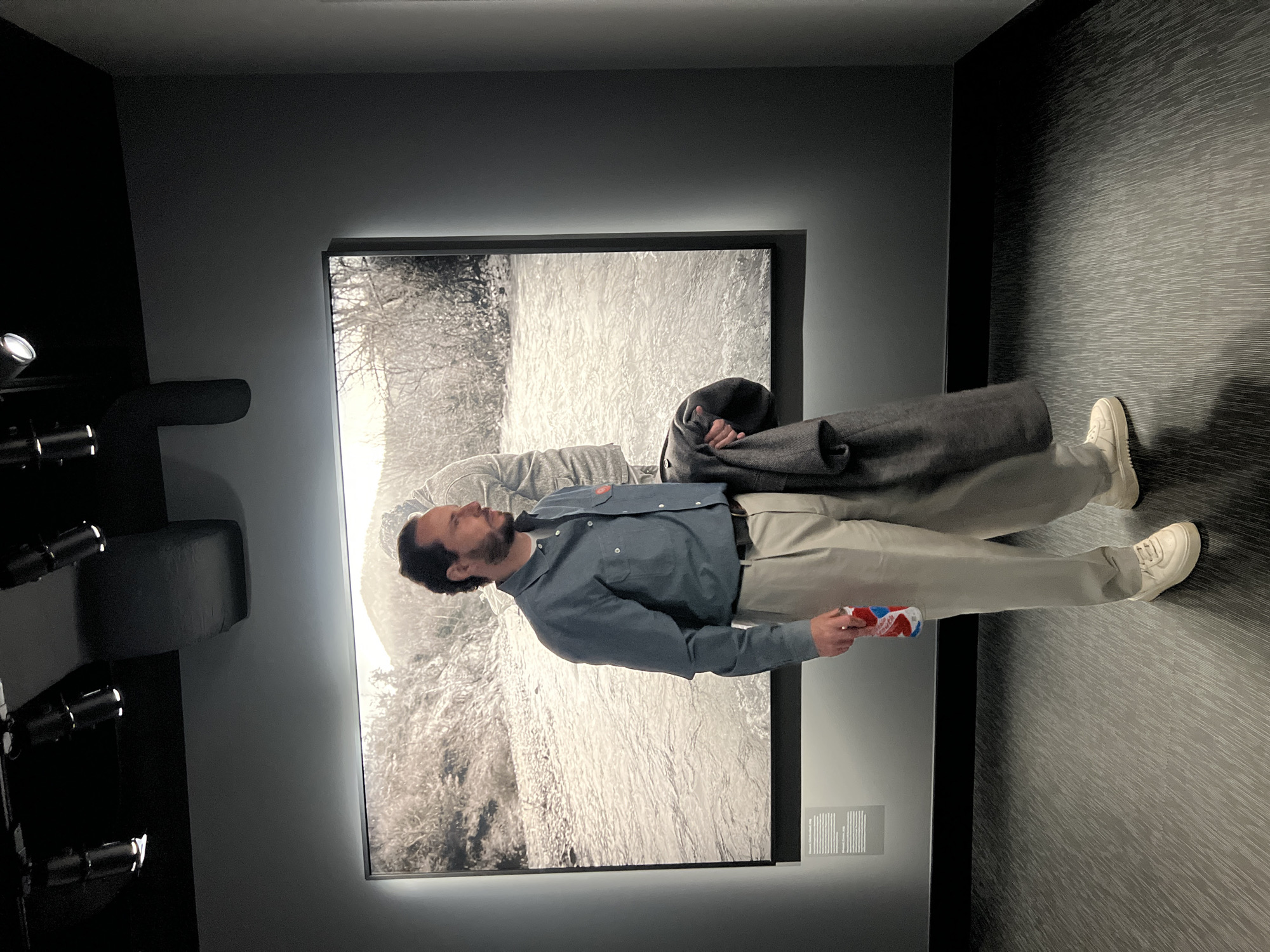 The image shows a man standing in a room in Berlin, Germany. He is wearing a blue shirt and beige pants and holding a colorful item in his right hand. He is standing in front of a large black-and-white artwork hanging on a gray wall. There is a gray coat draped over part of the artwork. To the left of the man and artwork is a black object that appears to be part of an exhibit. The floor is dark gray with a textured pattern.