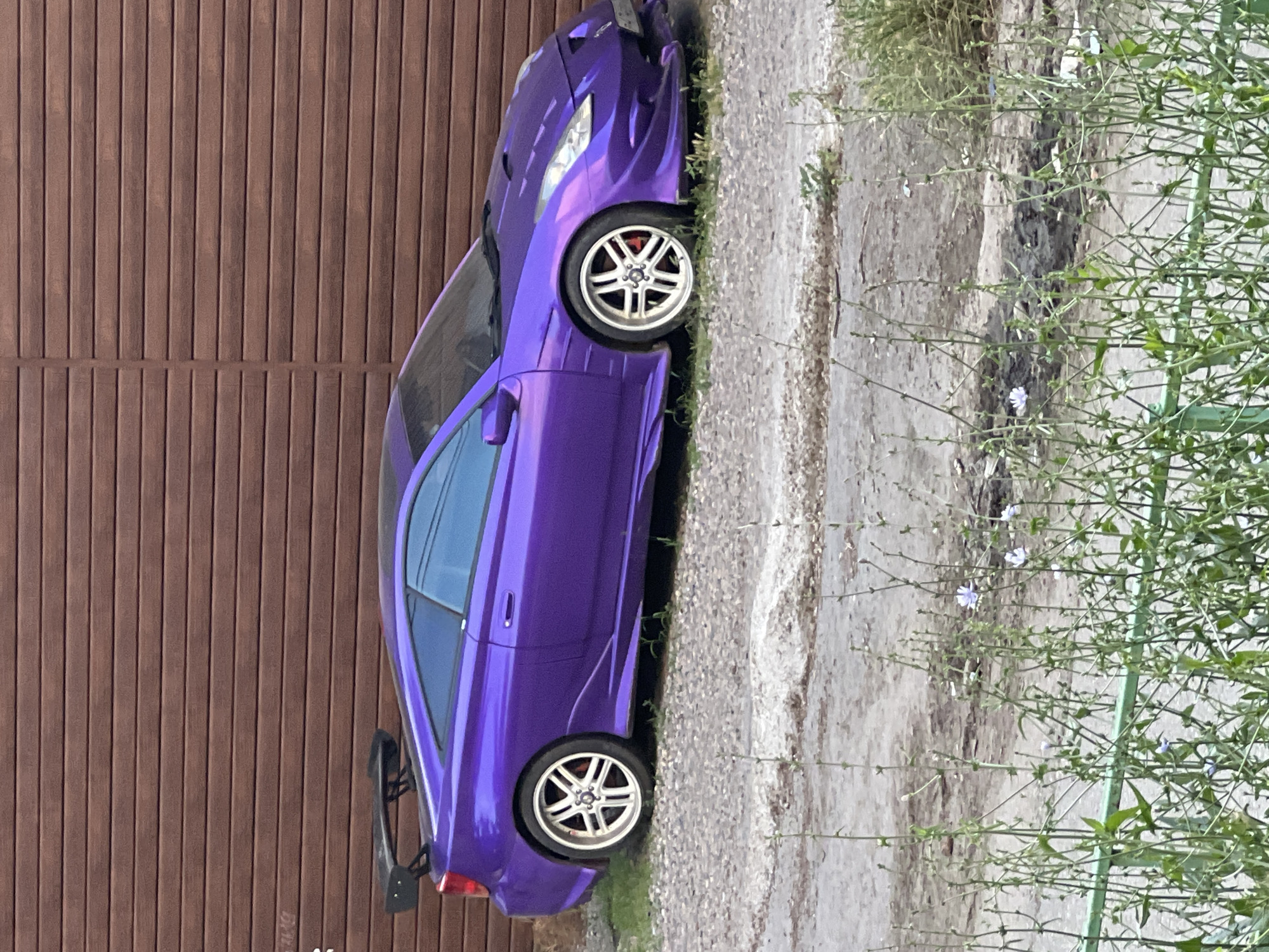 A modified purple Toyota Celica is parked on a gravel lot in front of a brown wooden fence. The car has a prominent rear spoiler, custom body kit, and aftermarket wheels. The scene is outdoors with some greenery and weeds in the foreground. The image appears to be taken in an urban or suburban setting.