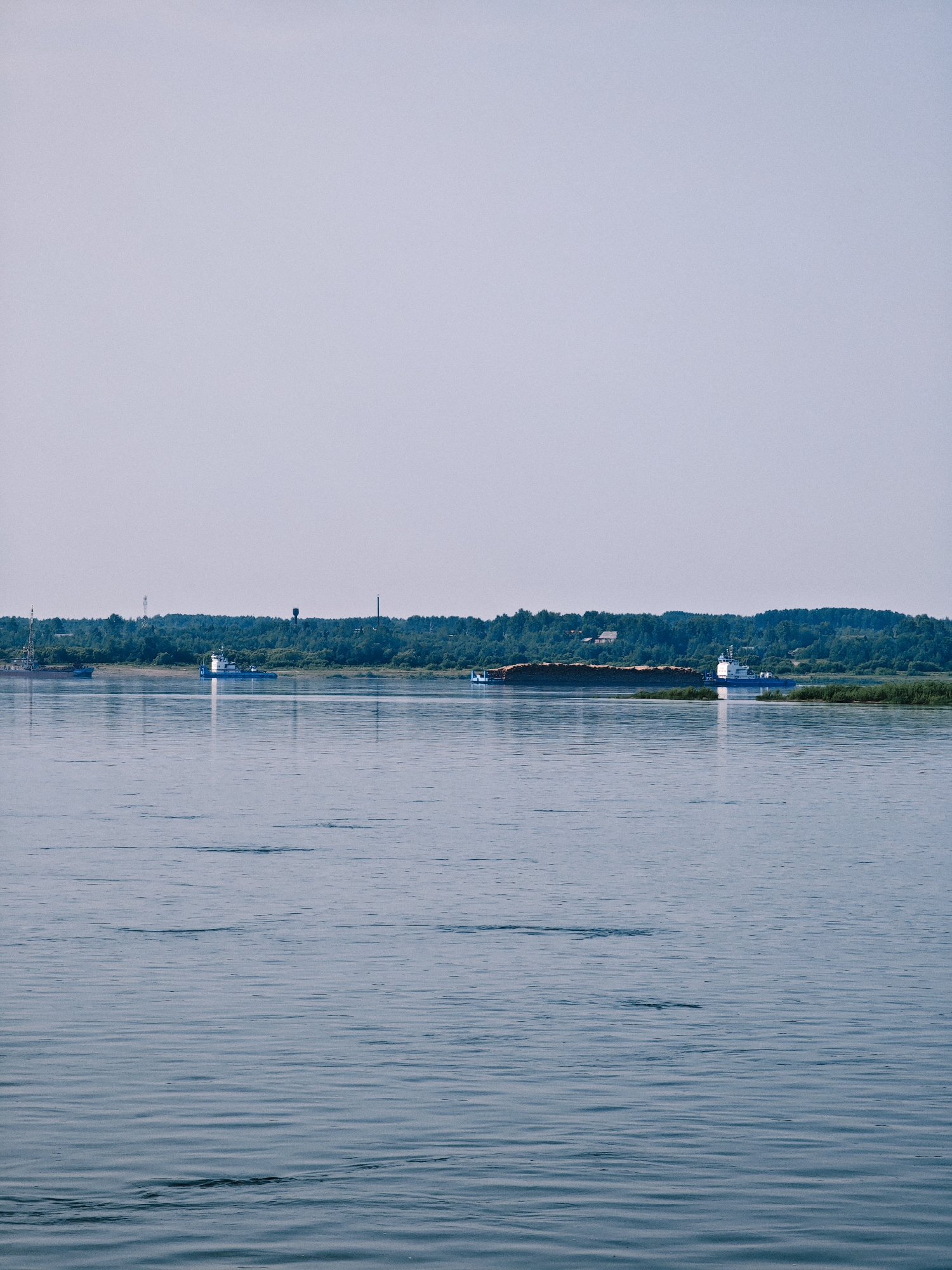 A wide, calm river dominates the foreground, reflecting the pale, hazy sky. Along the distant shore, a dense line of trees forms a dark green border against the horizon. Several boats are visible on the water. On the left, a barge with a crane is moored near the shore. Further to the right, a white tugboat is actively maneuvering. Another barge, heavily laden with what appears to be logs, is being pushed by a tugboat. The scene suggests a working river or port area. The city of Novonazimovo, Russia, is noted as the location.