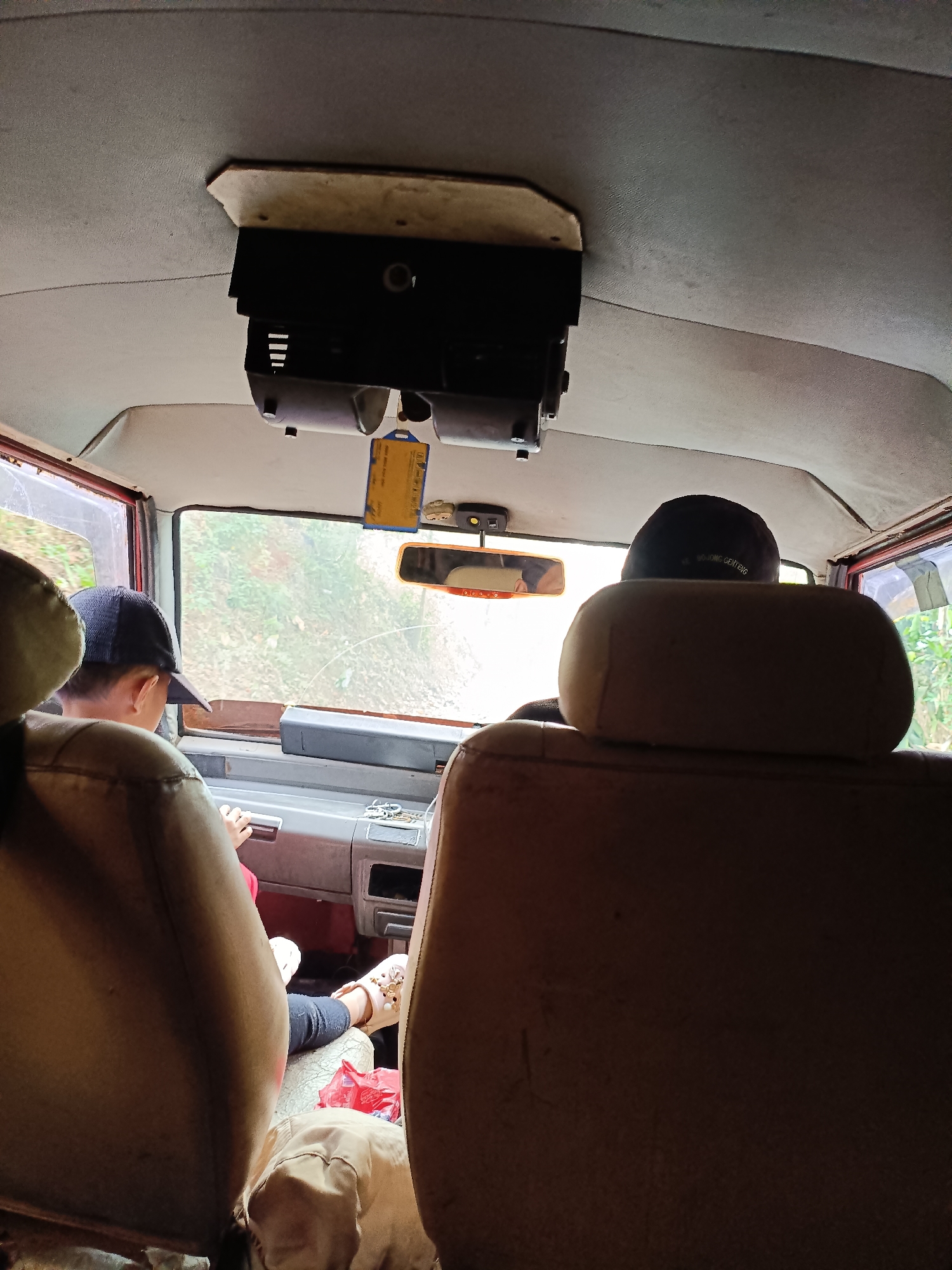 The image shows the interior of a vehicle, likely a van or jeep, with two individuals visible. In the foreground, facing away from the camera, are two headrests of seats. The view is directed towards the front of the vehicle, through the windshield.
A person wearing a dark baseball cap and a red shirt is seated in the driver's position, with their hands on the steering wheel or dashboard. They appear to be looking forward. The dashboard area is visible, with some controls and a rearview mirror.
To the right of the driver, another person is seated, also facing forward, with their back to the camera. They are wearing a dark cap that appears to have text on it, possibly "BOJONG GENTENG". The text on the cap is partially obscured.
The windshield shows an outdoor environment with green foliage and trees, suggesting the vehicle is on a road or path surrounded by nature. A yellow tag is hanging from the rearview mirror. The interior of the vehicle appears somewhat dated, with fabric-covered headliner and seats. The overall lighting suggests it is daytime.