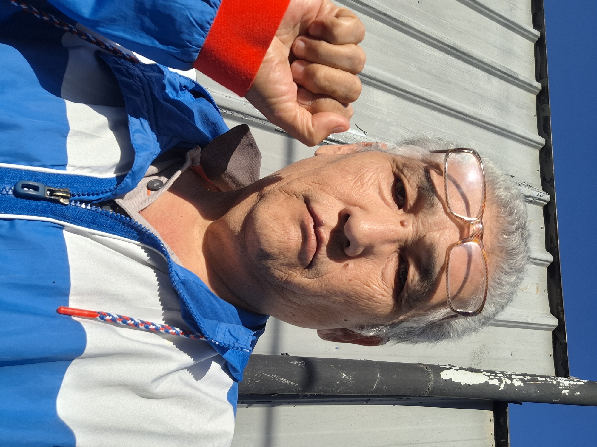 The image is a close-up selfie of an elderly East Asian male with graying hair, wearing glasses with brown frames. He has wrinkles on his face and is wearing a blue and white hooded jacket with red trim on the sleeve. His left fist is clenched and raised near his face. The background consists of a corrugated metal wall and a clear blue sky. A black pole with peeling white paint is visible to the right of the man. The scene appears to be outdoors, possibly in an urban environment. There is no specific indication of the city of Almaty, Kazakhstan within the image itself.