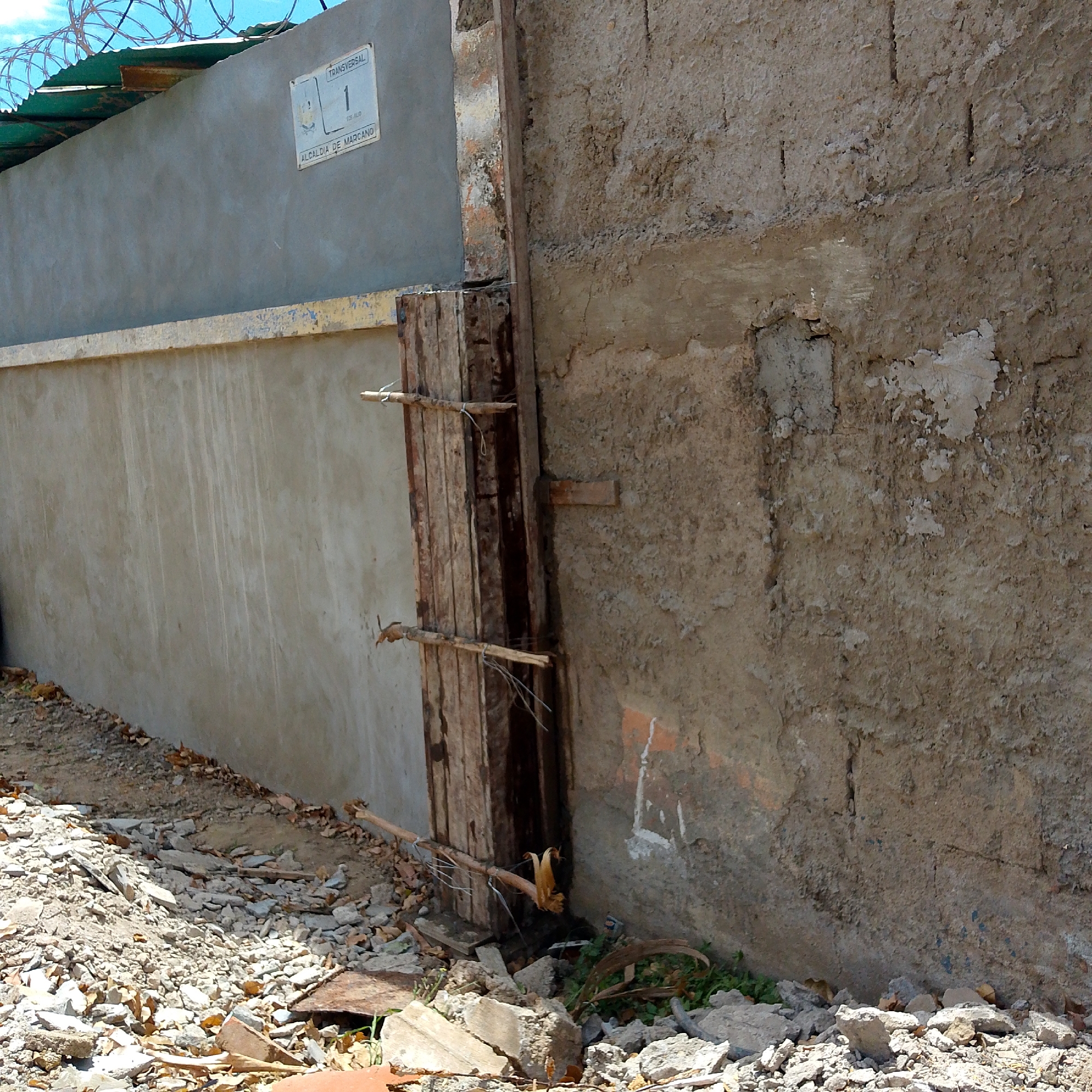 The image depicts a damaged exterior wall in a possible urban environment. Two walls are visible, constructed of different materials. The wall on the left appears to be made of concrete and is painted gray. A sign is affixed to this wall that says "TRANSVERSAL 1" and "ALCALDIA DE MARCANO", which suggests this is the city of Altagracia in Venezuela. The wall on the right is rougher, with a texture indicative of mud brick construction and is in a state of disrepair, with sections patched or missing. A wooden structure is positioned between the two walls, propping up the mud brick wall. Debris litters the ground, including stones and broken pieces of concrete or brick, which could suggest recent construction, demolition, or a lack of maintenance. There are no people visible in the frame.