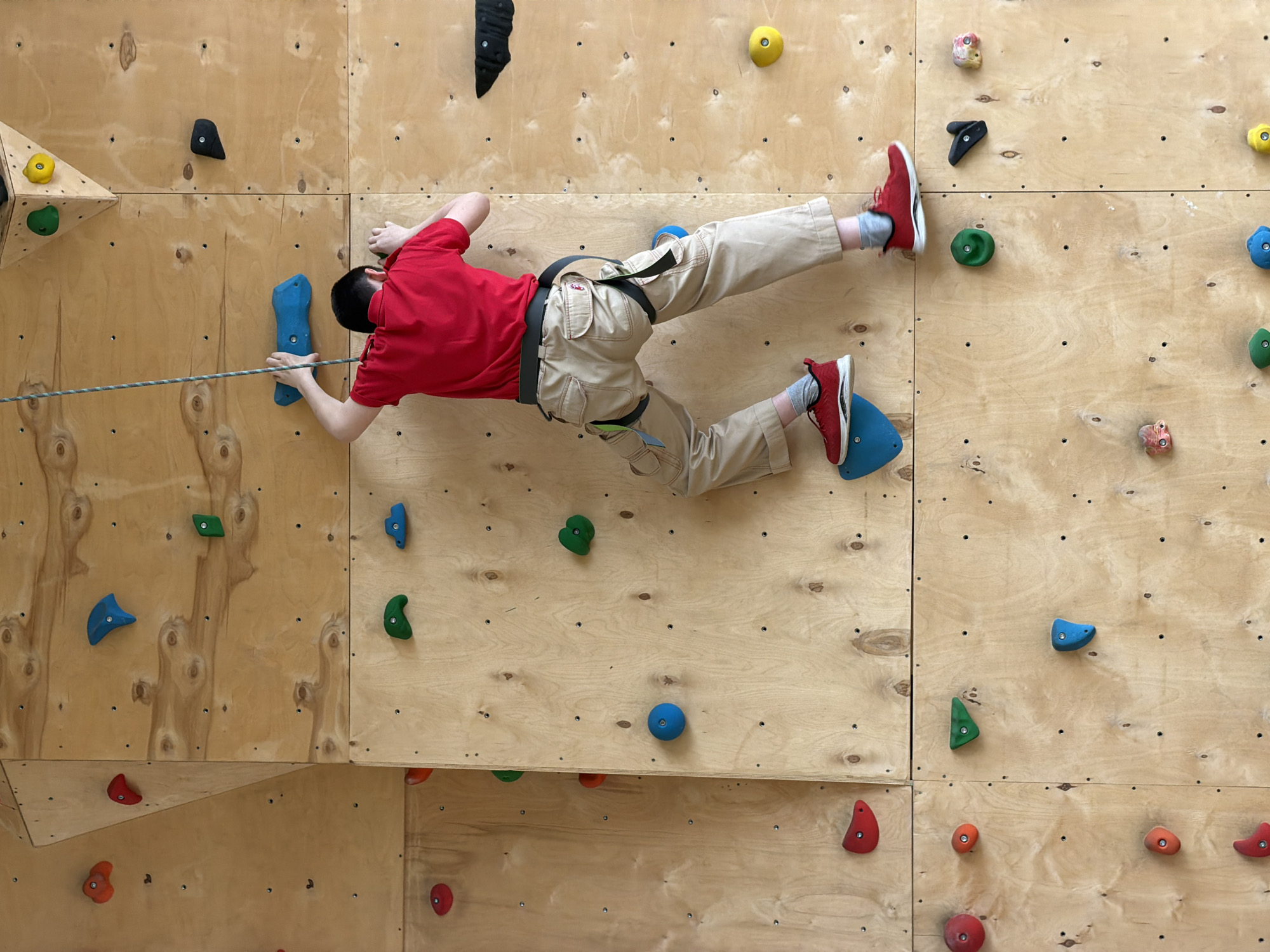 An indoor climbing wall in Izhevsk, Russia, is depicted with a male youth or young adult actively climbing or traversing its surface. The individual, dressed in a red t-shirt, khaki cargo pants, red sneakers, and a black climbing harness, is positioned horizontally across the light-colored plywood panels of the wall. His left hand grips a prominent blue hold, while his feet are placed on other blue and red holds, demonstrating a dynamic climbing posture. A green and white patterned rope extends across the wall and passes through his hands.
The climbing wall is constructed from numerous rectangular plywood sections, fitted with a variety of colorful climbing holds in shades of yellow, green, blue, black, red, orange, and mottled pink/white. These holds are bolted directly into the wooden panels. On the left side of the frame, a three-dimensional wooden feature, resembling a triangular corner or overhang, adds to the complexity of the climbing terrain. The lighting in the indoor facility appears bright and even. No text is visible in the image, and the climber's face is obscured, making it impossible to discern specific emotions, though his body language suggests focus and exertion.
