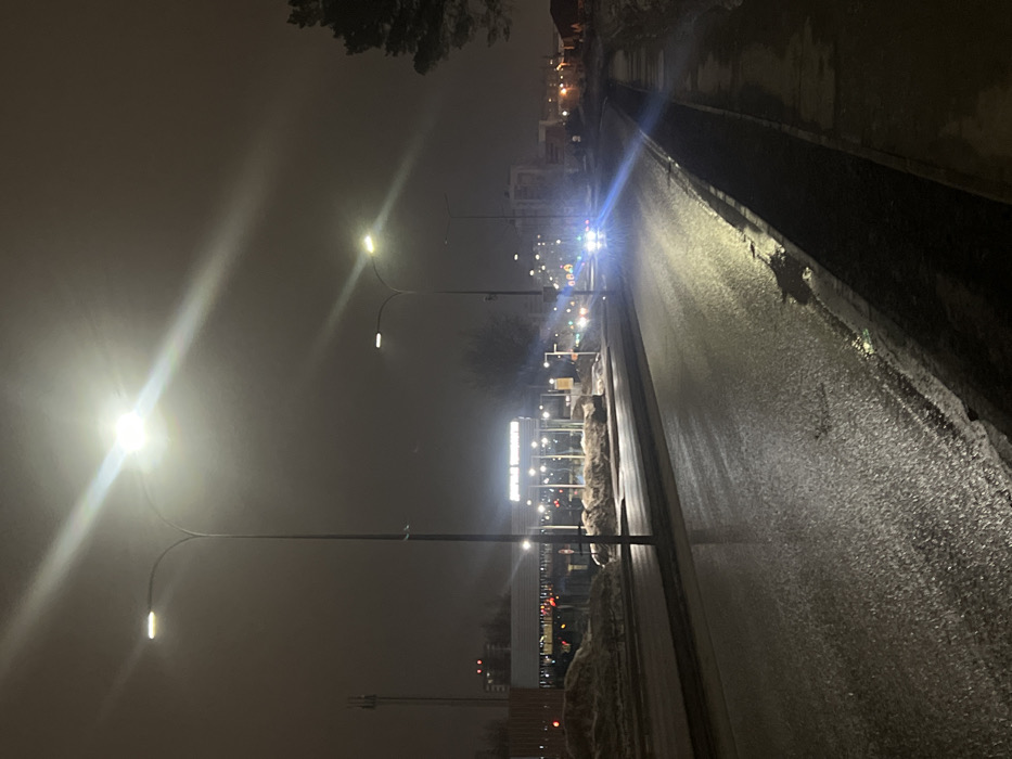 The image captures a nighttime urban street scene in Yelabuga, Russia, specifically looking east along Botkina Street near building 3A. The environment features an overcast sky, likely with fog or haze, and wet ground conditions suggesting recent precipitation or melted snow.
A two-lane asphalt road, with one lane designated for each direction, extends into the distance. The road surface is dark, reflective, and visibly wet. Along the right side of the road, a wet sidewalk parallels the thoroughfare, exhibiting accumulated patches of dirty snow and slush along its edge. A large conifer tree, partially dusted with snow, is discernible on the far right.
On the left side of the road, across an unpaved median containing mounds of dirty snow, stands a brightly illuminated commercial building. Its rooftop sign, partially obscured by light glare, displays Cyrillic text including "ДОБРОЦЕН," identifying it as a supermarket. Further left, a tall communication tower is visible against the dark sky.
Multiple streetlights illuminate the scene, emitting strong, diffused light that creates visible halos and beams due to atmospheric conditions. One prominent streetlight in the immediate left foreground casts a bright glow on the wet road surface. In the distance, intense headlights from an approaching vehicle are visible on the right lane of the road, producing significant glare. No individuals are present within the frame. Other urban structures are dimly visible further down the street.