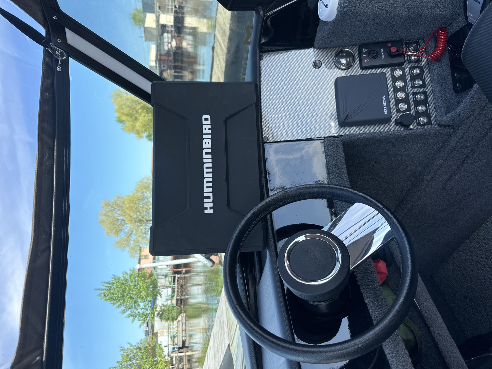 The image shows the interior of a boat cockpit. A black Humminbird fishfinder is prominent on the left. A black steering wheel with chrome accents is in the foreground. The dashboard features various Honda-branded controls and gauges. The view through the windshield reveals a waterfront scene with buildings and trees, reflective of the sky. Based on the visual cues and the identified brand associations (Honda marine controls), the boat appears to be located on the Kama River in Chaikovsky, Russia.