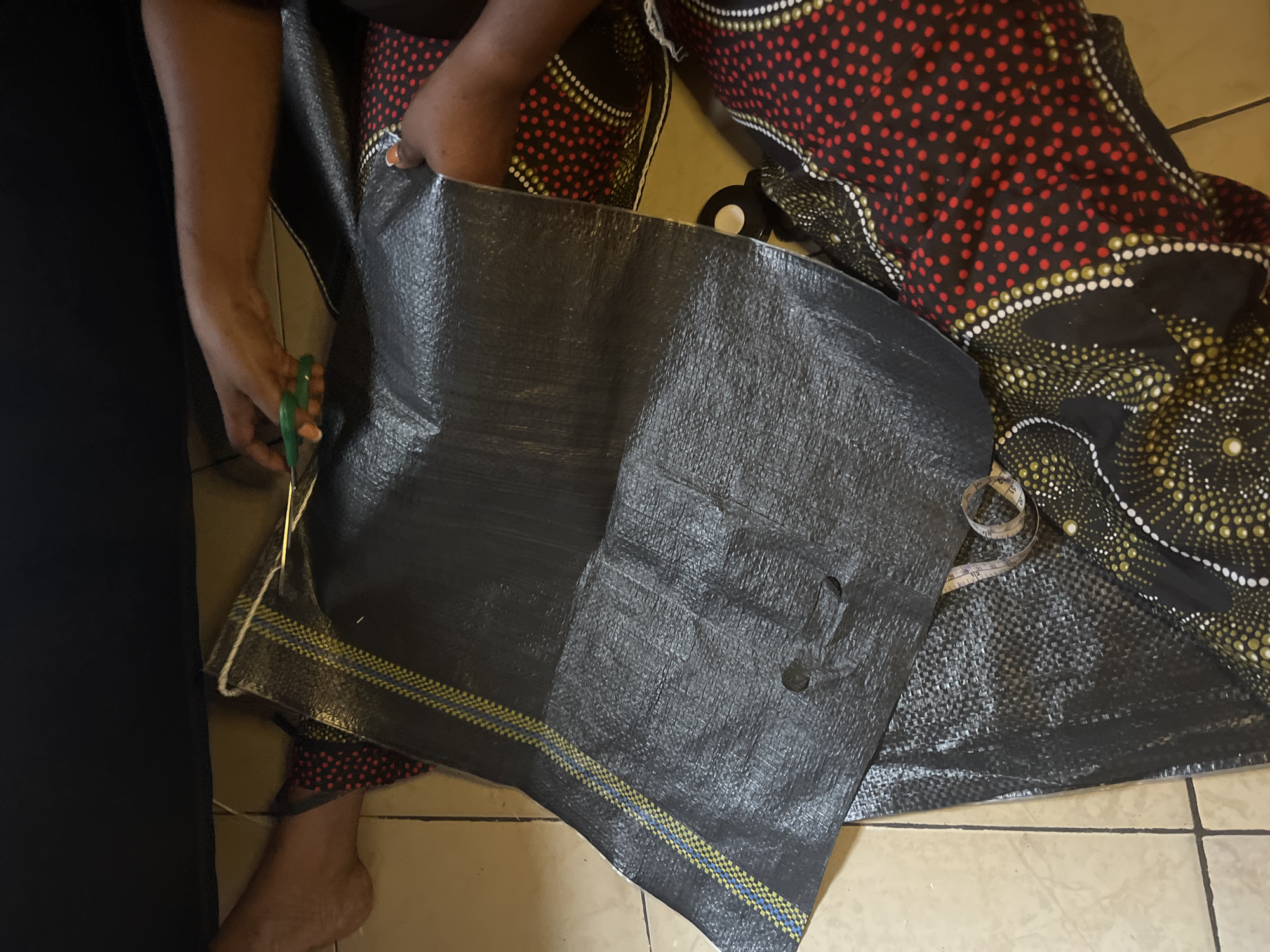 In Badagry, Nigeria, an individual is using green-handled scissors to cut a large, black woven plastic bag. The bag has yellow and blue stripes along the bottom edge. The person is wearing a patterned fabric skirt or pants with red polka dots and a black fabric with gold and white circles. A measuring tape lies nearby on a similar black woven bag that lies under the patterned fabric. The background shows a tiled floor and part of a dark object.