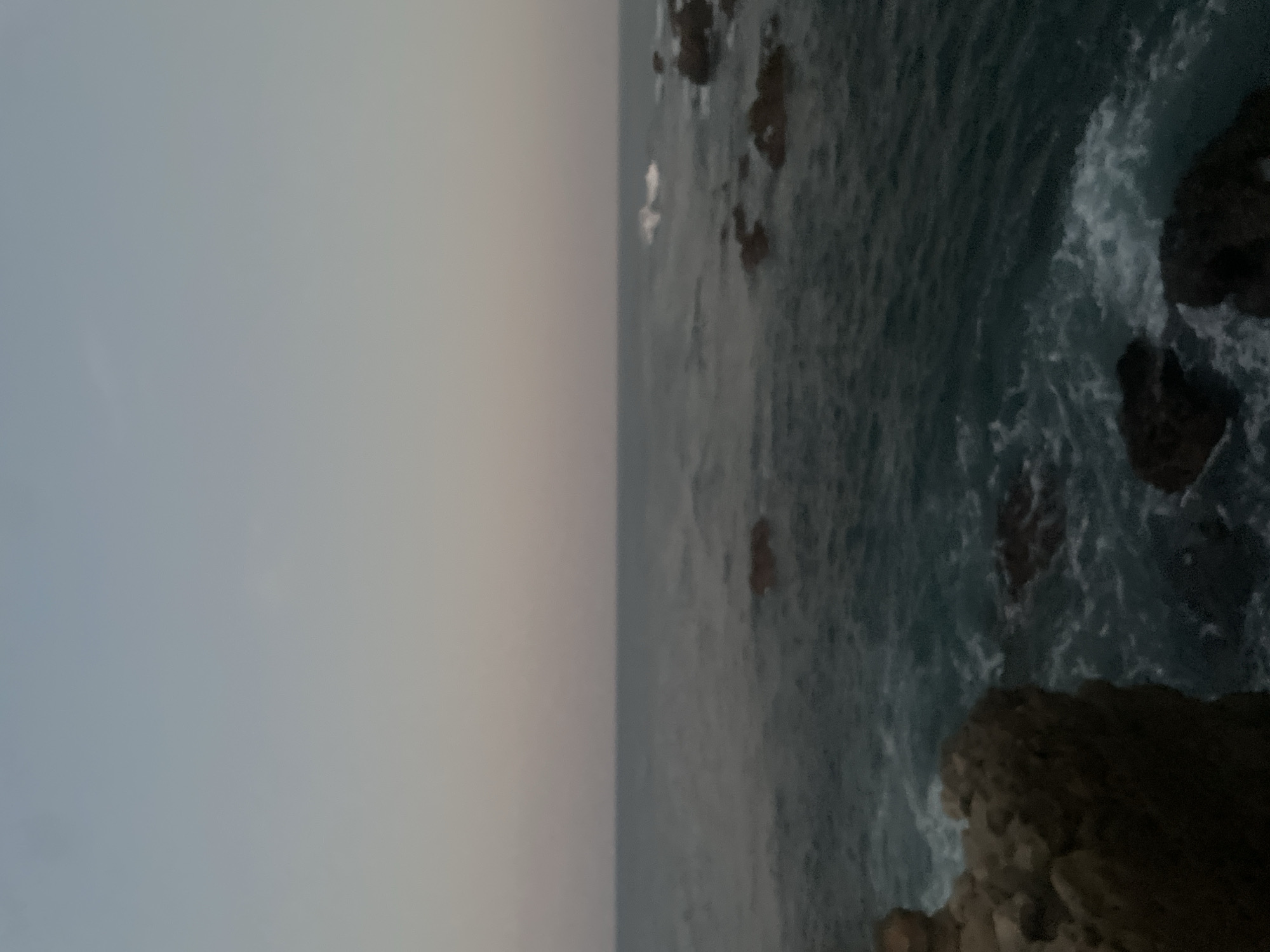 The image captures a seascape in Puerto Escondido, Mexico. The sky is overcast, creating a diffused lighting condition. The ocean is turbulent, with visible waves crashing against rocky outcroppings. There are no people visible in the frame. The focus is on the natural environment, specifically the contrast between the sky and the sea.