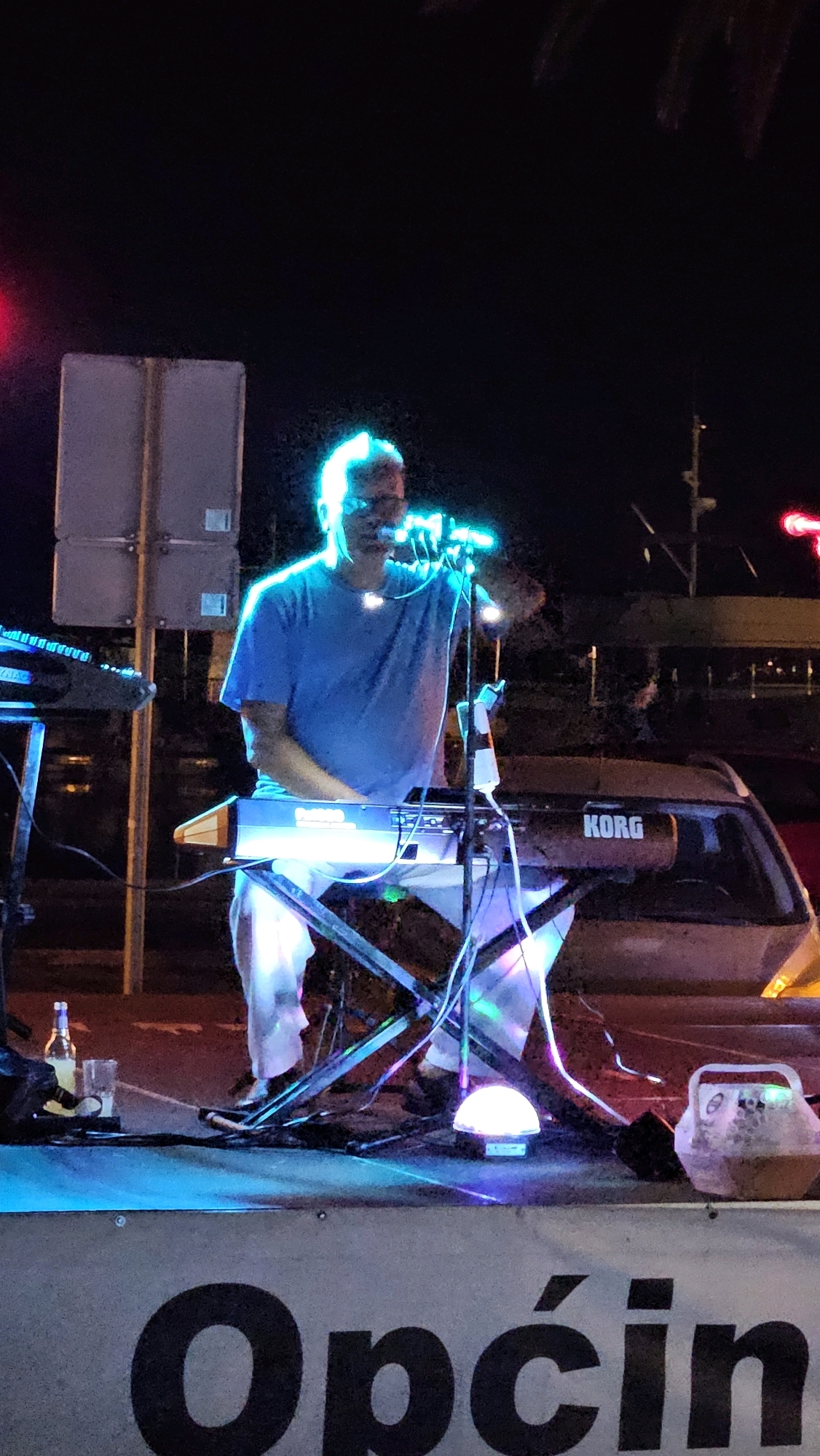 A man plays a Korg keyboard on a stage at night in Općina Tučepi, Croatia. The man is wearing a blue t-shirt and white pants and is illuminated by blue lights. He is holding a microphone. A keyboard stand and various other musical equipment are visible on the stage. In the background, there are palm trees and buildings. The words "Općin" are visible on a banner at the bottom of the image.