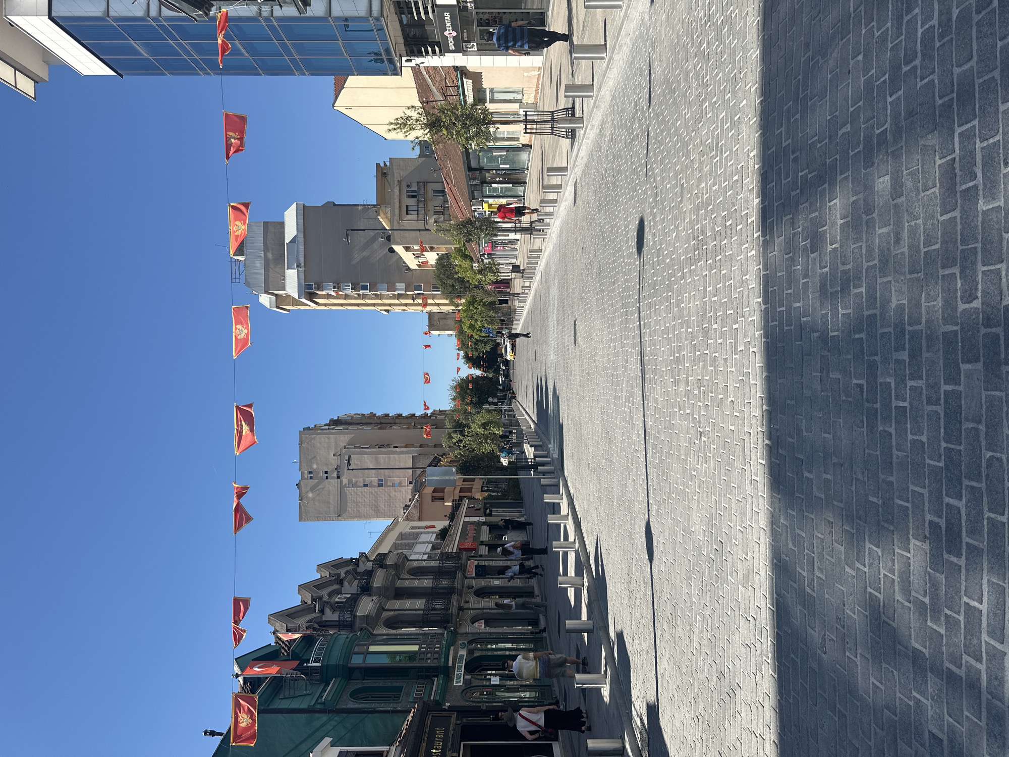 The image shows a street in Podgorica, Montenegro, lined with buildings of varying heights and architectural styles. Red flags are strung across the street. The street is paved with gray bricks. A row of metal bollards is placed along one side. People are walking on the street and sitting outside of shops and restaurants. The sky is clear and blue. A shadow is cast diagonally across the brick pavement.