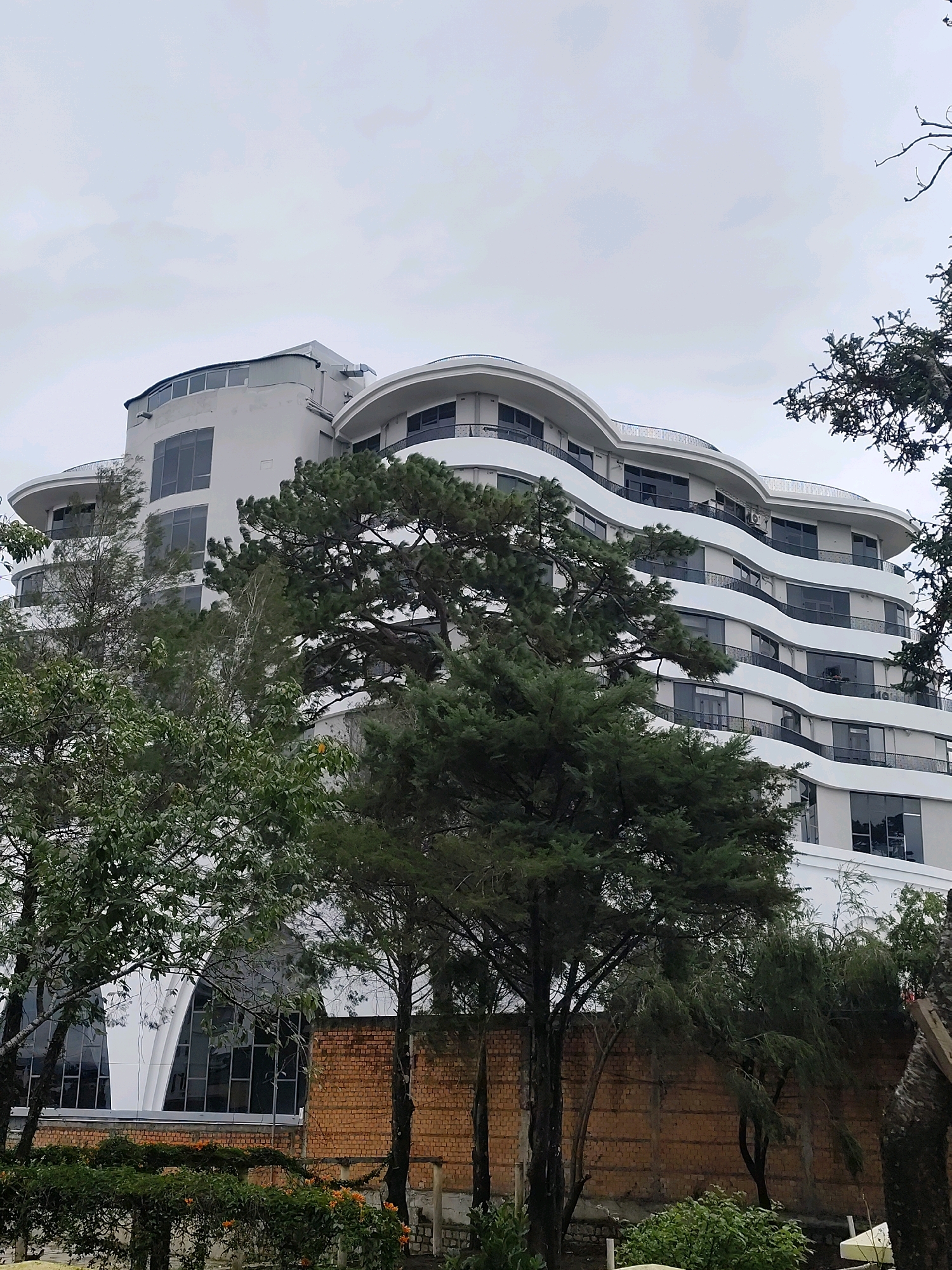 The image captures a large, white, multi-story building with a distinctive wave-like architectural design in Da Lat, Vietnam. The building features curved balconies with dark railings and numerous large windows. In the foreground, several lush green trees, including pine trees, partially obscure the view of the building. Behind the trees, a brick wall is visible. The sky is overcast and appears to be a pale gray. The overall scene suggests a modern building set within a natural, slightly misty environment. This building is the Dalat Palace Hotel.