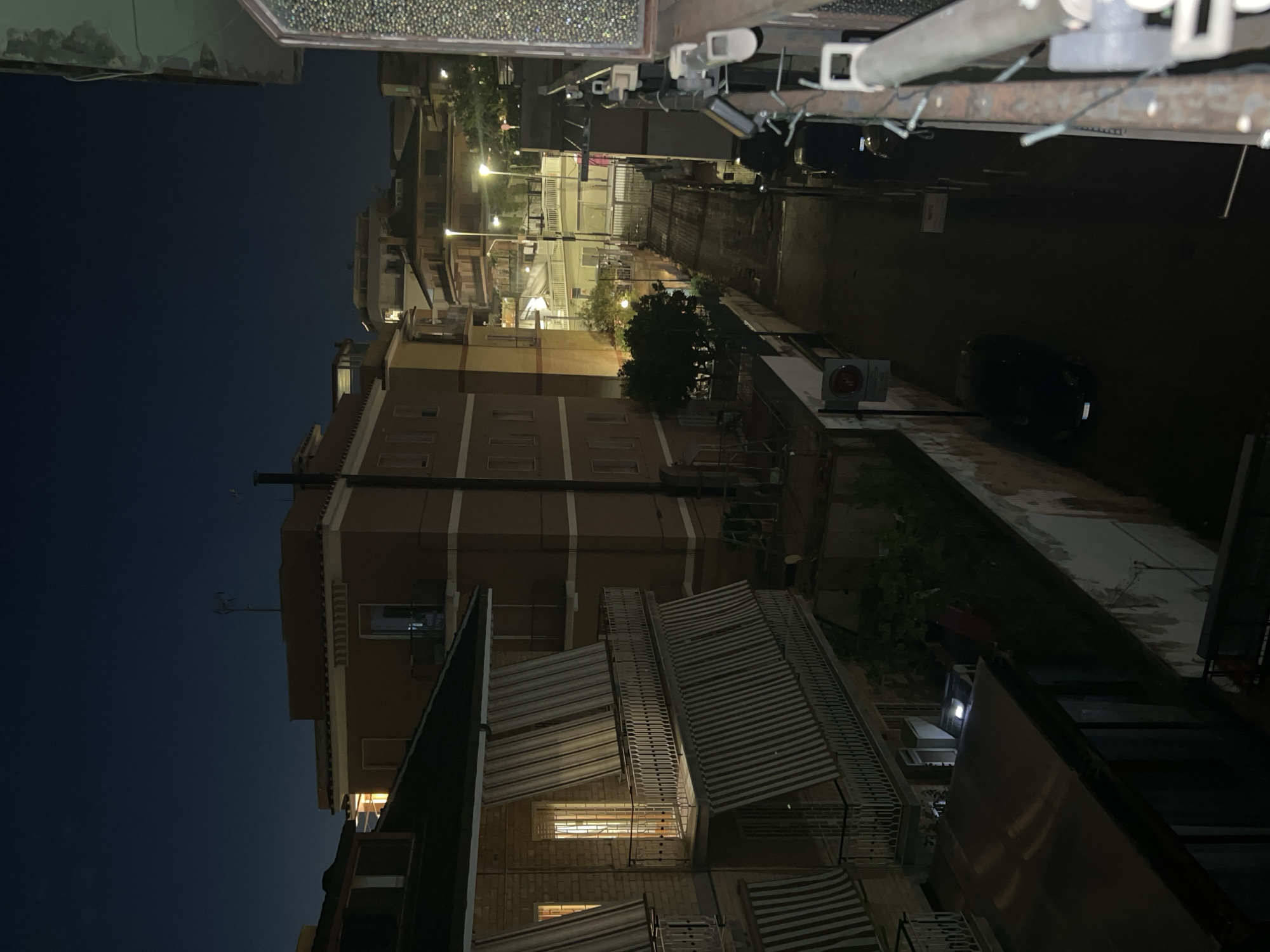 The image depicts a narrow street in Rome, Italy, at night. The scene is dominated by apartment buildings on either side of the street, illuminated by streetlights and interior lights from windows. One building on the left has balconies with striped awnings and ornate railings. The street itself is paved and appears wet, reflecting the ambient light. Two parked cars are visible on the street. The sky is dark blue, indicating nighttime. The overall impression is one of a residential urban environment after dark.