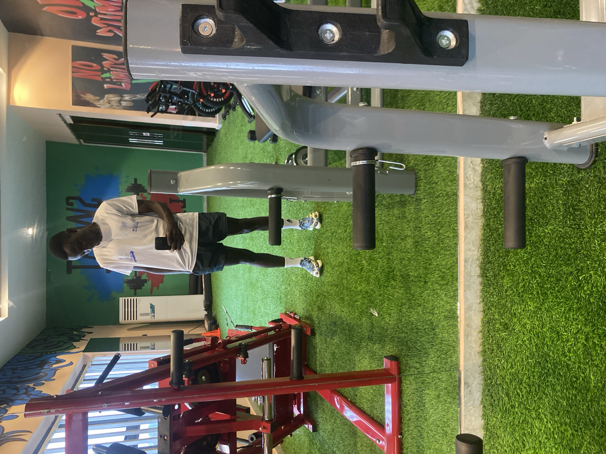 This is a full-body, slightly overhead, mirror-shot photograph taken inside a gym. A young Black man, dressed in a white t-shirt and dark shorts, stands in the center of the frame, looking down at his phone, which he holds in his right hand. He is wearing white socks and athletic shoes. The gym has a bright, artificial turf floor. To the left, a large, red weight machine with chrome accents is visible. To the right, a gray weight machine is partially in the foreground, with the man's legs and feet in the background. The walls of the gym are painted with colorful murals, including blue and white abstract designs on the left and motivational text and imagery on the right. An air conditioning unit is visible on the back wall. The gym is located in Barden Yana, Nigeria.