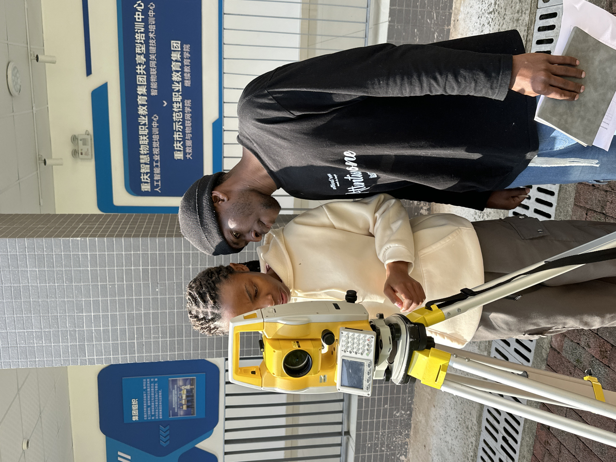 Two individuals are observed near a surveying instrument set up on a tripod. One person is operating the device, while the other appears to be observing or instructing, and holding a notebook. The location is identifiable as Jiangjin, China, based on signage indicating "Chongqing Smart IoT Vocational Education Group Sharing Training Center" and "Chongqing Demonstration Vocational Education Group." The environment suggests an urban setting, with tiled walls and street grates visible.