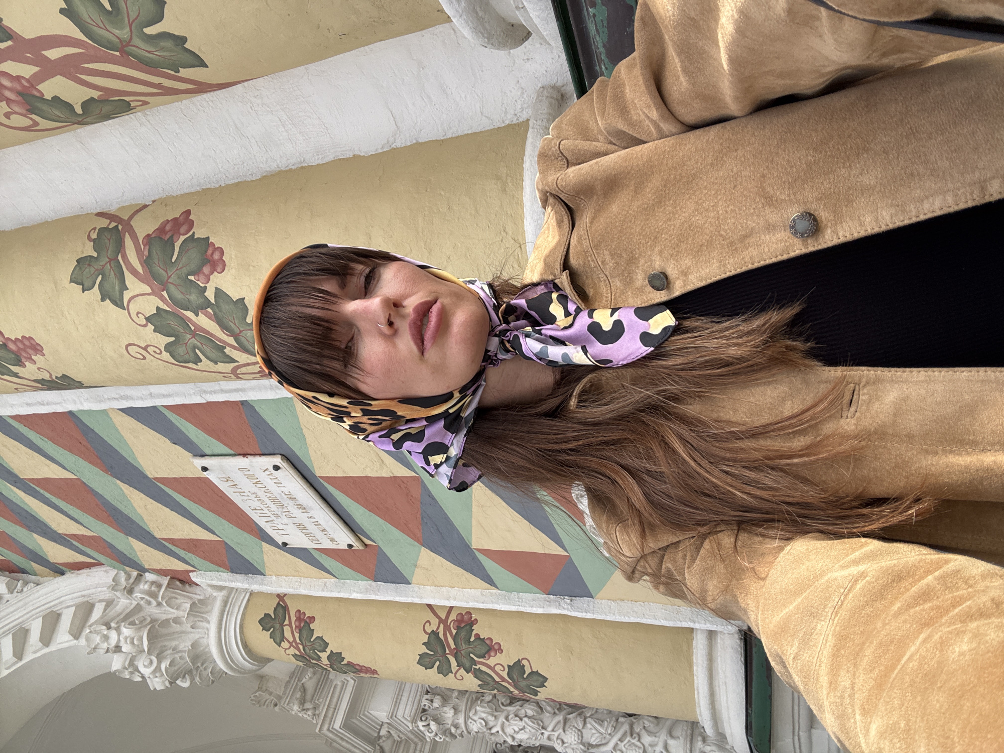 A woman wearing a brown suede jacket and a colorful headscarf sits in front of a building with a multi-colored striped facade and decorative columns. A sign above her head reads "ТРАПЕЗНАЯ ЦЕРКОВЬ СЕРГИЯ РАДОНЕЖСКОГО СООРУЖЕНА В 1682-1692 ГОДАХ", indicating that this is the refectory church of Sergius of Radonezh, built between 1682 and 1692, located in the city of Sergiyev Posad, Russia. The background features a wall adorned with painted grapevines.