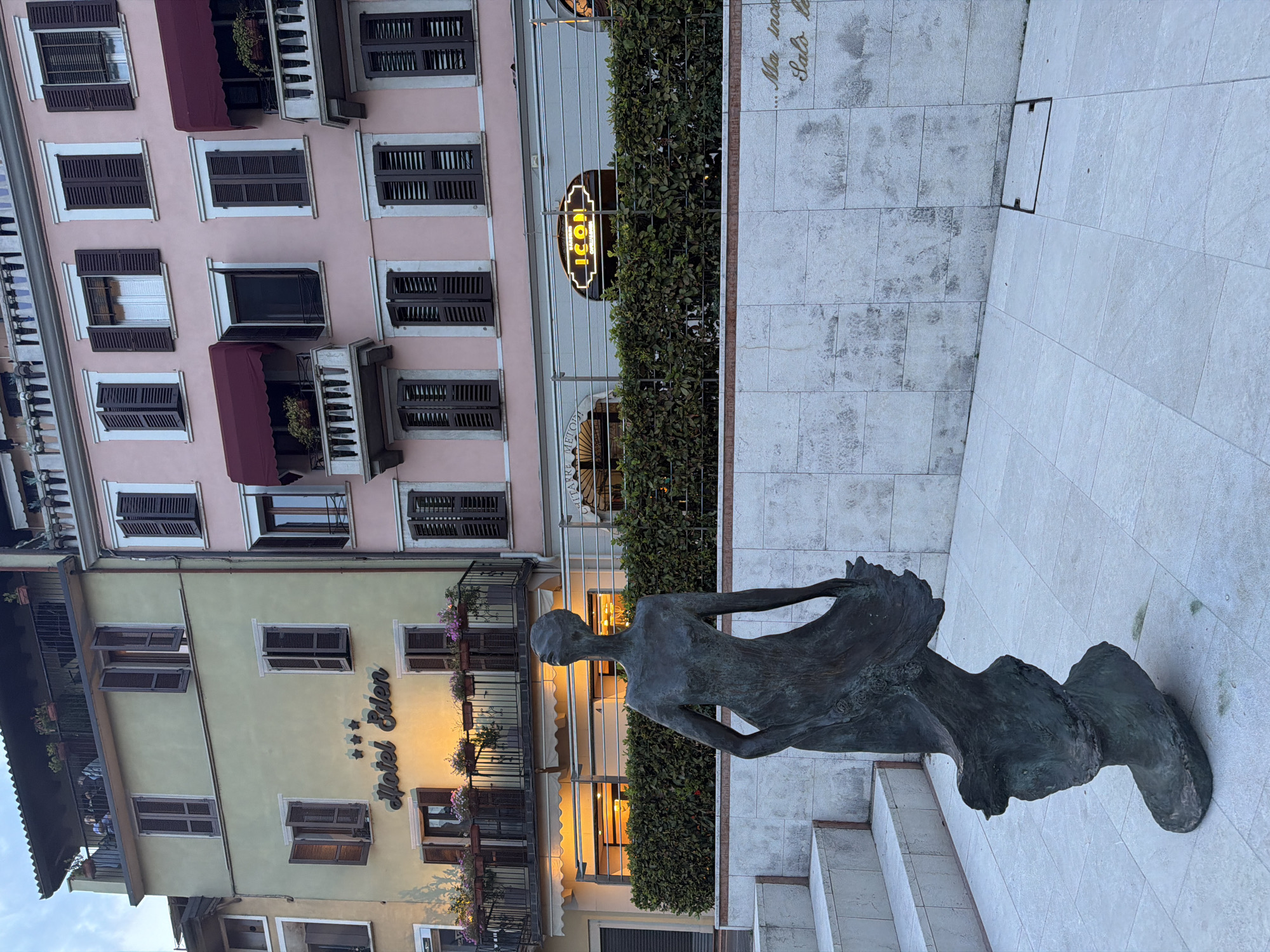 In Salò, Italy, a bronze sculpture of a woman in a flowing dress stands on a tiled plaza in front of a building with a pale yellow facade on the left and a pink facade on the right. The sculpture's head is tilted back, and its arms are outstretched. The yellow building has a sign that reads "Hotel Eden" with three stars above it. Several windows with dark brown shutters are visible on both buildings, some open and some closed. Balconies with potted plants adorn the upper floors. Below the balconies, a row of green bushes lines the facade. A neon sign with the word "ICON" is illuminated above a business on the pink building. The ground is paved with large, light-colored tiles. In the background, the sky is overcast. To the left of the sculpture, there are tiered steps made of the same light-colored tiles. On the right side of the wall behind the sculpture, there is faint, cursive text.