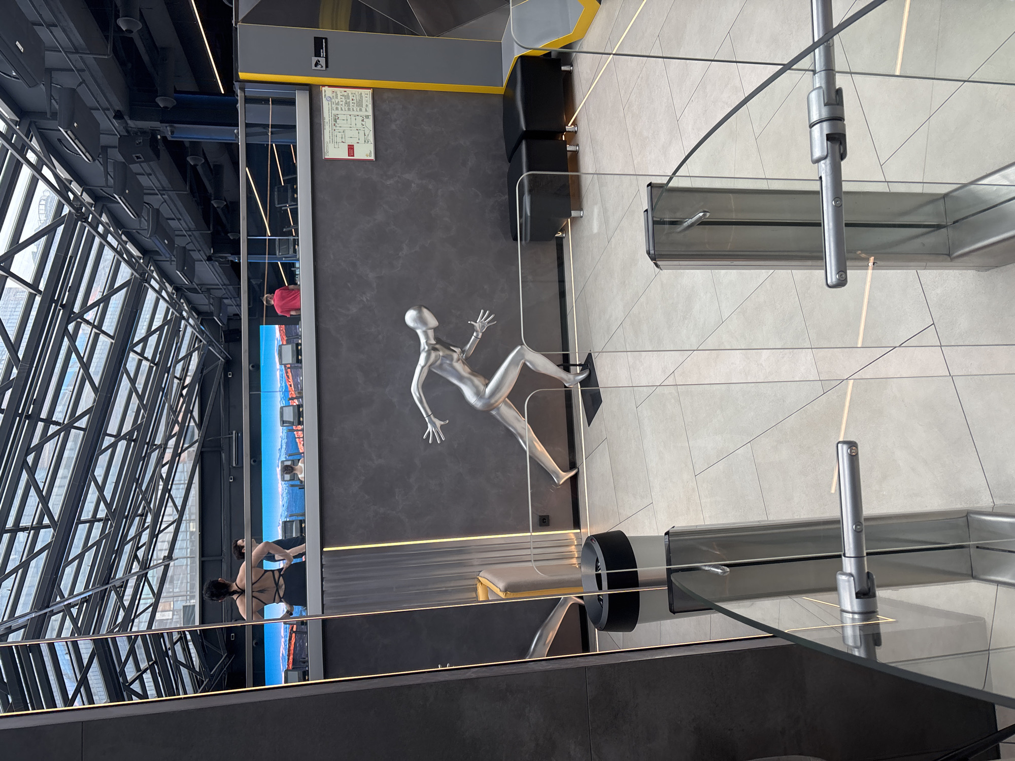 The image shows an interior view of a high-rise building in Murino, Russia. The distinctive architectural structure of the skyscraper is seen through a glass wall with visible steel support beams and a cityscape beyond. Inside, there is a dark gray stone floor, with a silver mannequin lying on its side. There are some black square seats on the dark gray stone floor next to the walls. Adjacent is a waist-high glass partition with steel supports, creating a barrier or viewing area. In the background, a screen displays an image of a beach scene with people, and another person, dressed in red, is visible in the distance. A woman with a complex strap top is standing in front of the screen, near the glass wall. A building plan diagram is on the wall near the seats. The image captures the blend of modern architecture and interior design within the Murino high-rise, with a focus on the spatial arrangement and the juxtaposition of art, design, and architecture.