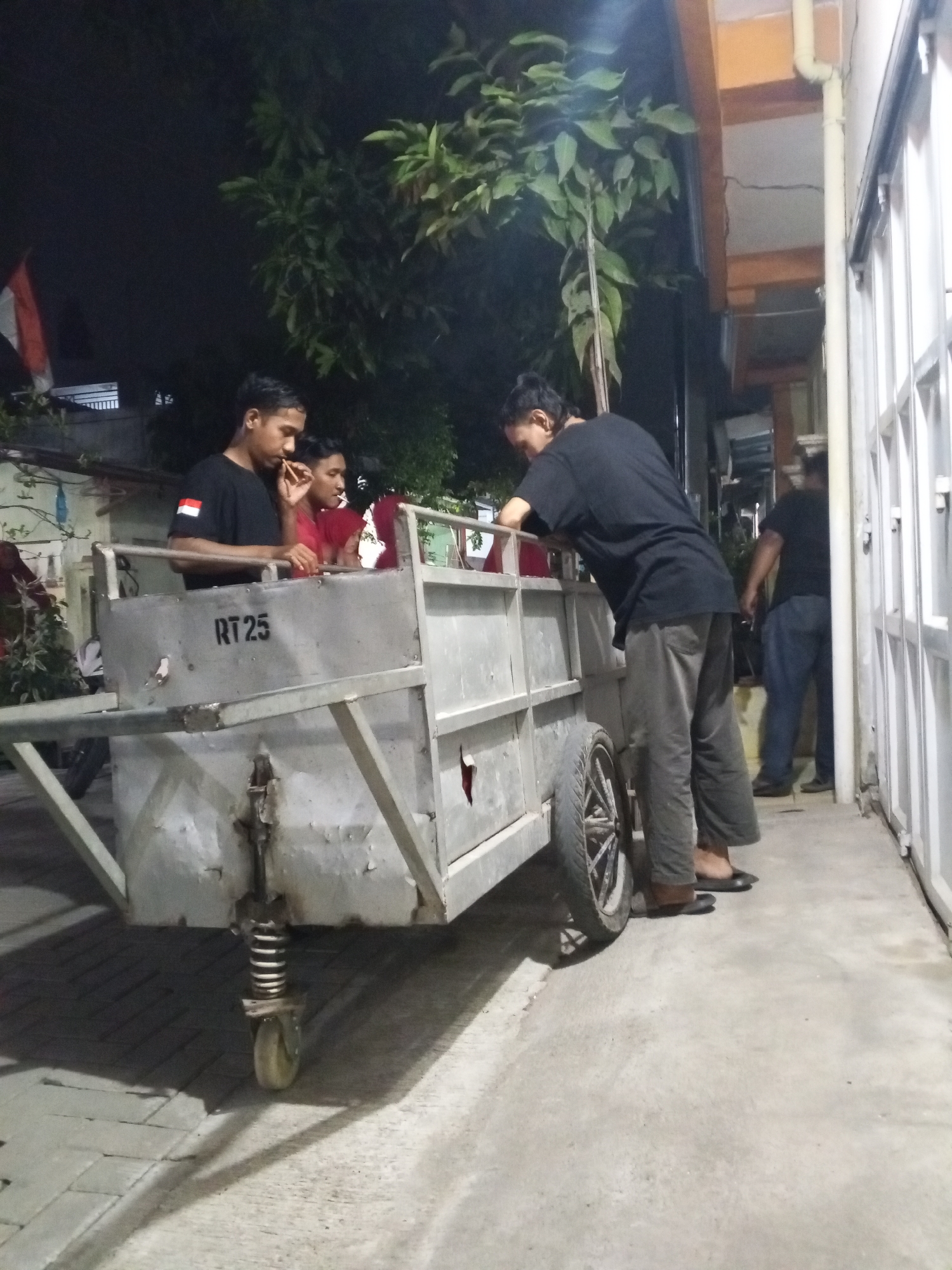 The image depicts an outdoor scene at night, possibly in the city of Ciruas, Indonesia, given the context. In the foreground, a man in a black t-shirt and grey pants is leaning on a metal cart with two wheels. He appears to be holding something up to his mouth. To his left, two young men are standing near the cart, one wearing a black t-shirt and the other a red t-shirt. The man in the red t-shirt also appears to be holding something to his mouth. The cart has "RT 25" stenciled on its side. In the background, another man in a black t-shirt and blue jeans is walking away from the camera. The environment suggests a residential or commercial area with a building facade and some vegetation visible. The lighting is dim, indicating it is nighttime.