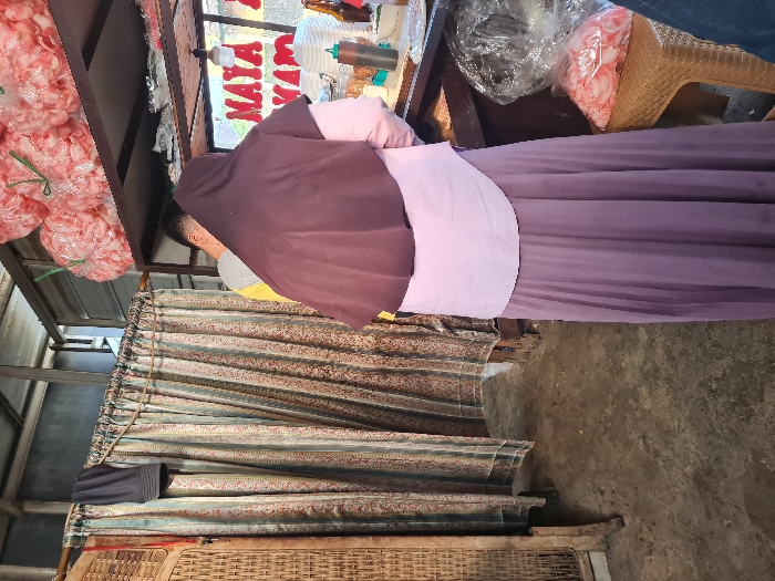 A woman in a purple hijab and a light pink sash stands at a food stall. Bags of pink snacks hang above. The stall has a faded sign. A woven basket and a patterned curtain are visible below. The ground appears to be concrete. Part of a wicker chair sits beside the woman.